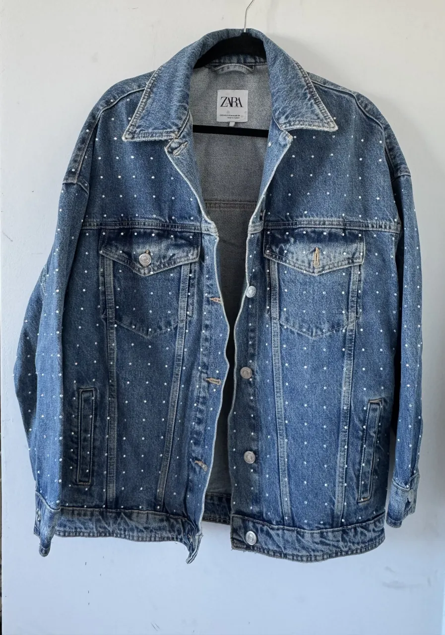 Campera de jean oversize azul con brillos distribuidas por toda la prenda. Tiene cuello solapa, bolsillos con tapa en el frente y botones metálicos.
