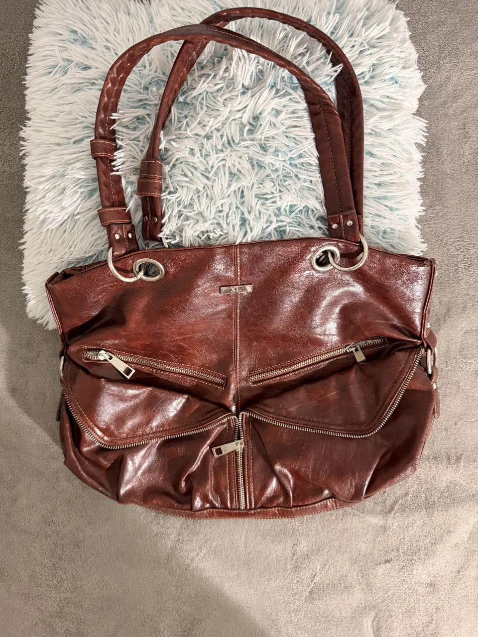 Bolso de cuero marrón oscuro, con múltiples bolsillos frontales con cierre y un bolsillo trasero con cierre. Las asas son de cuero trenzado y tiene detalles metálicos plateados. Ideal para el día a día o para llevar tus esenciales con estilo.