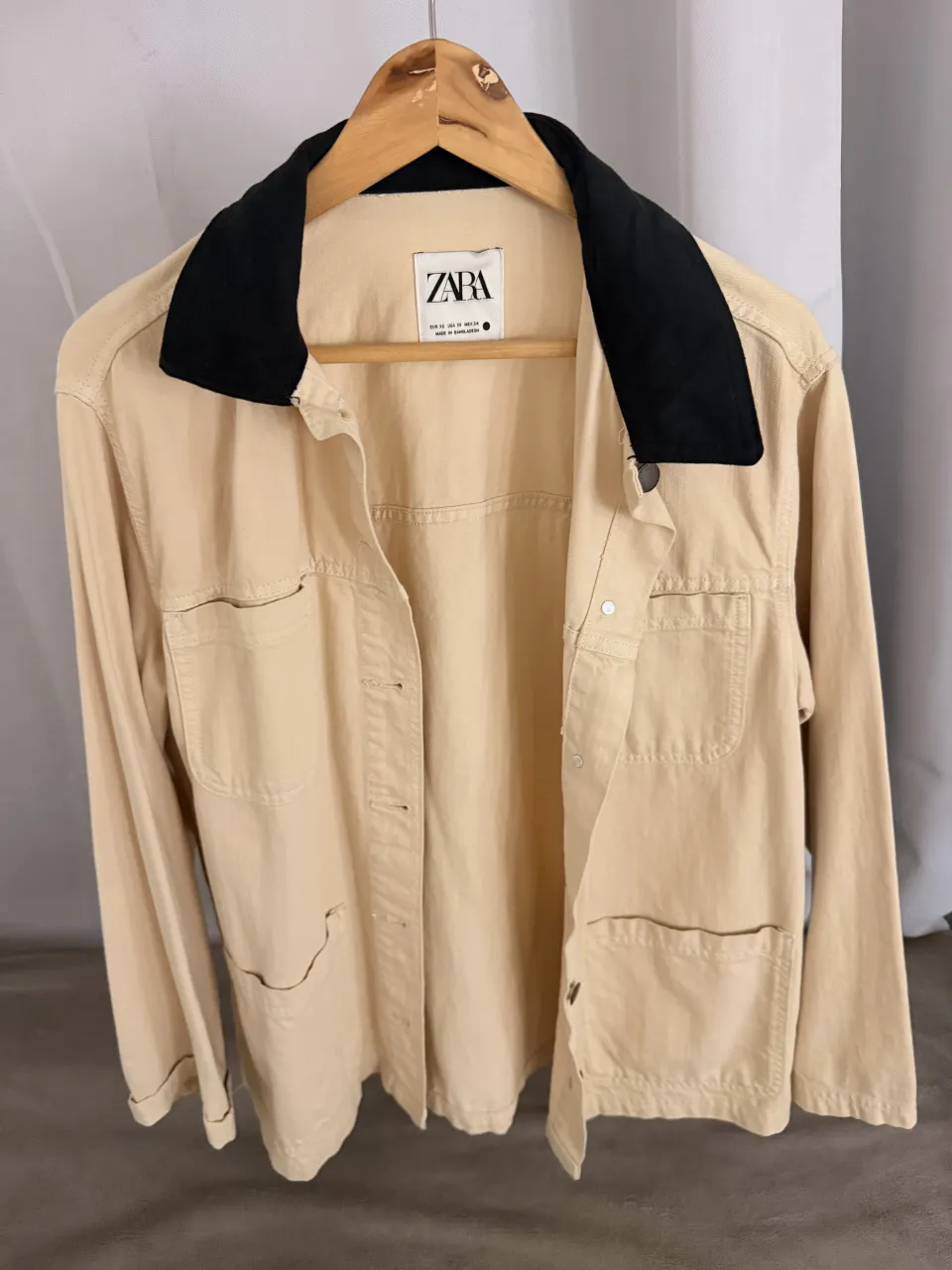 Campera de gabardina color tiza con cuello negro. Tiene bolsillos frontales y se abrocha con botones.