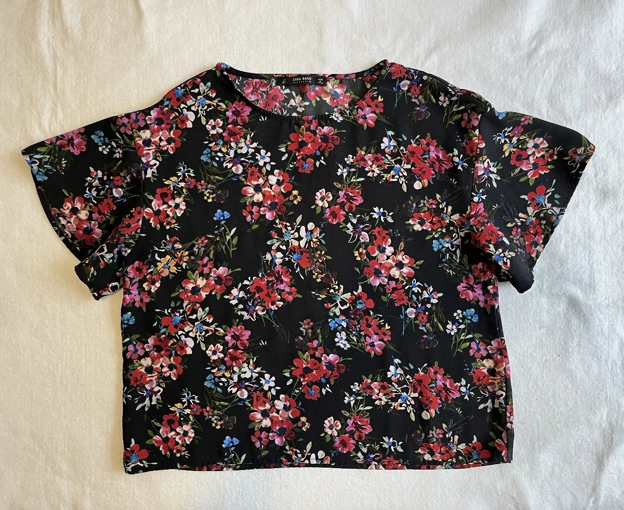 Blusa de mujer con estampado floral en tonos rojos, rosas y azules sobre fondo negro. Corte holgado y mangas cortas amplias. Ideal para un look casual y fresco.