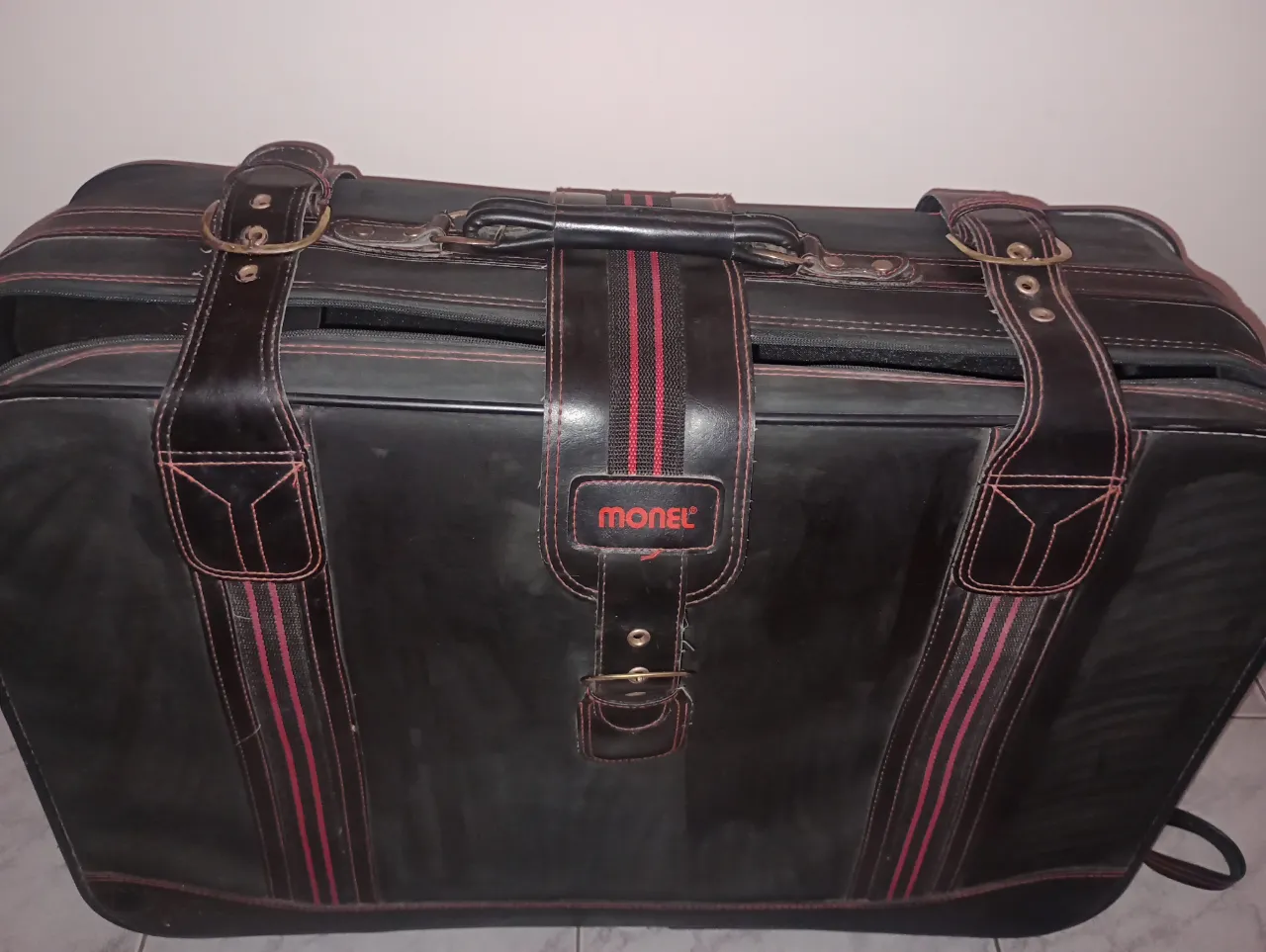 Valija vintage de cuero negra con detalles en rojo y negro. Ideal para viajes o como accesorio de moda con un toque retro. Posee correas de sujeción y hebillas.