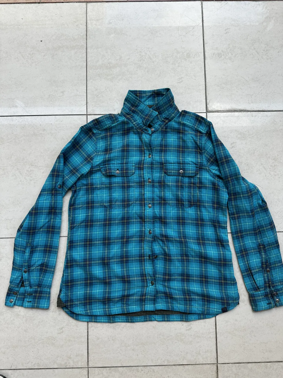 Camisa a cuadros de manga larga en tonos de azul y negro. Posee dos bolsillos en el frente y botones para cerrar.