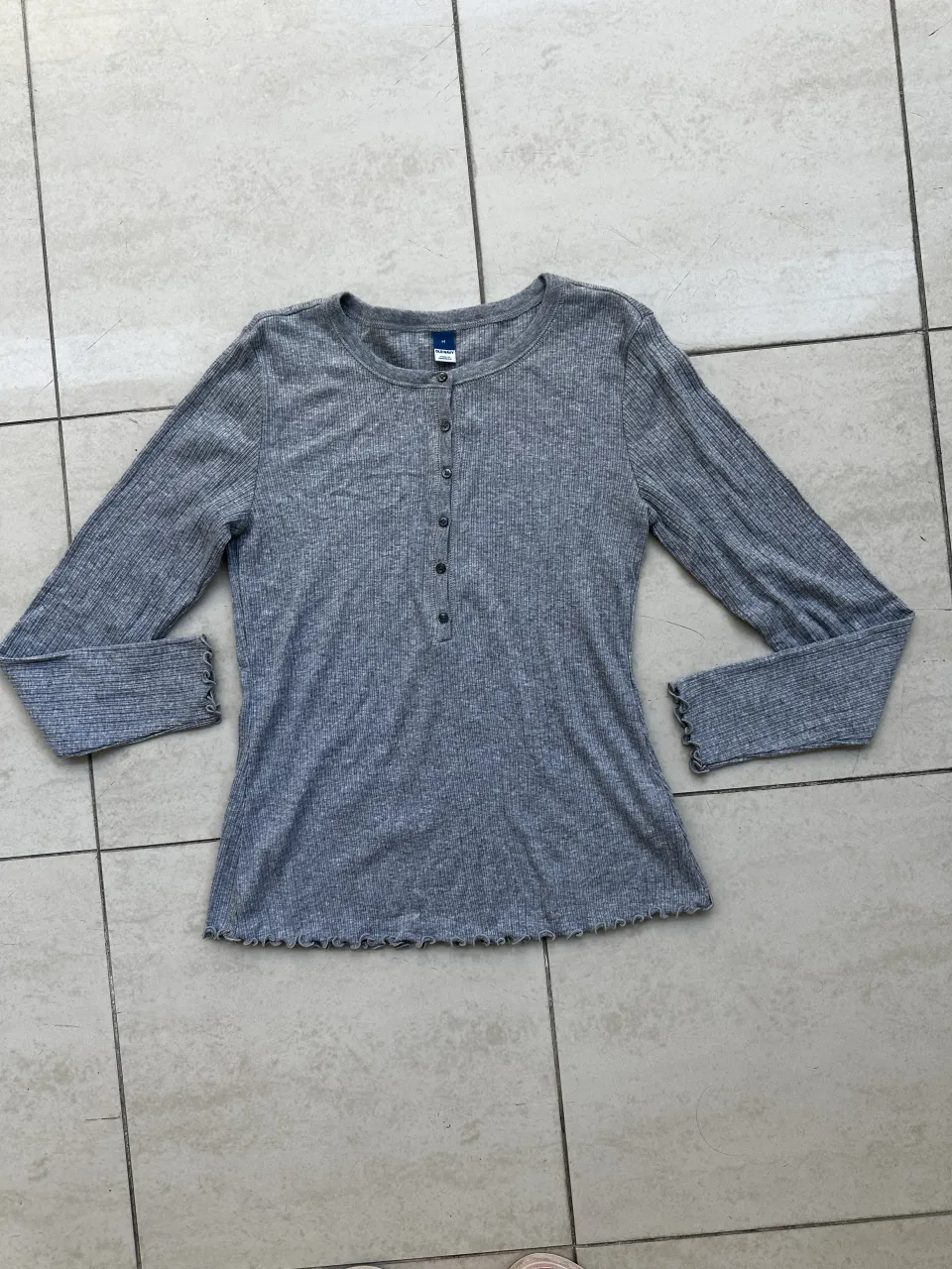 Remera gris de manga larga con cuello redondo y botones en la parte superior. Los puños y el ruedo tienen un detalle de volados.