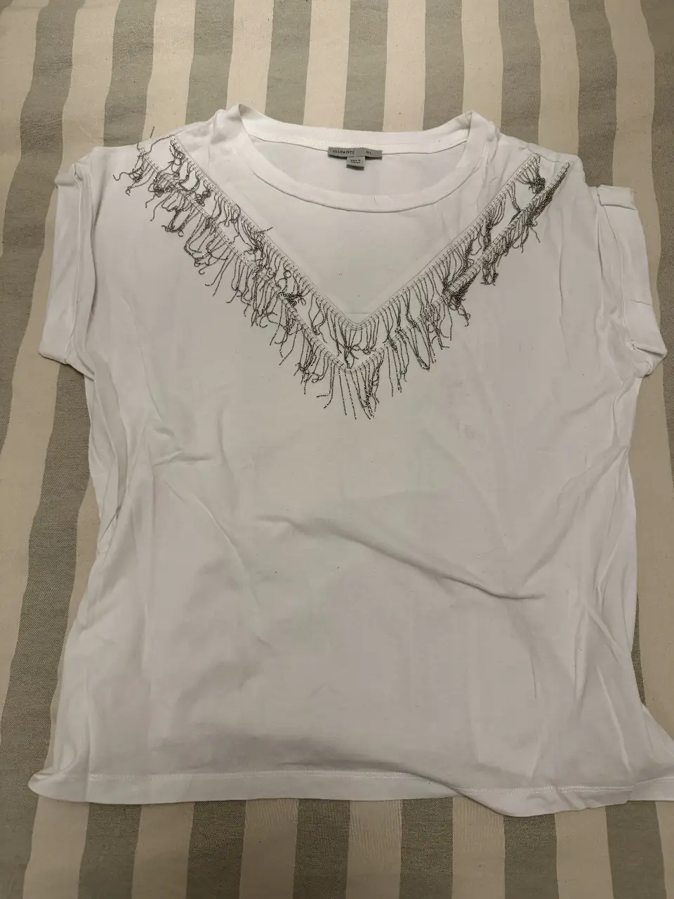 Remera blanca de mujer, con cuello redondo y detalle de flecos plateados en el escote. Talle M/L. Ideal para un look casual y canchero.