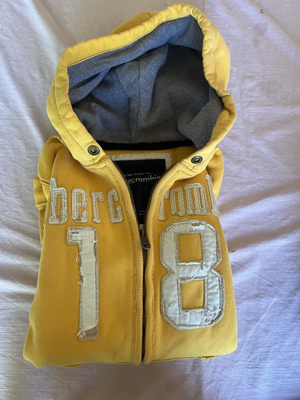 Buzo amarillo con capucha y cierre completo. Tiene apliques de letras blancas que forman "abercrombie" y el número "18" en el frente. En la manga tiene bordado "NY-10". talle L de niños pero es un S de mujer