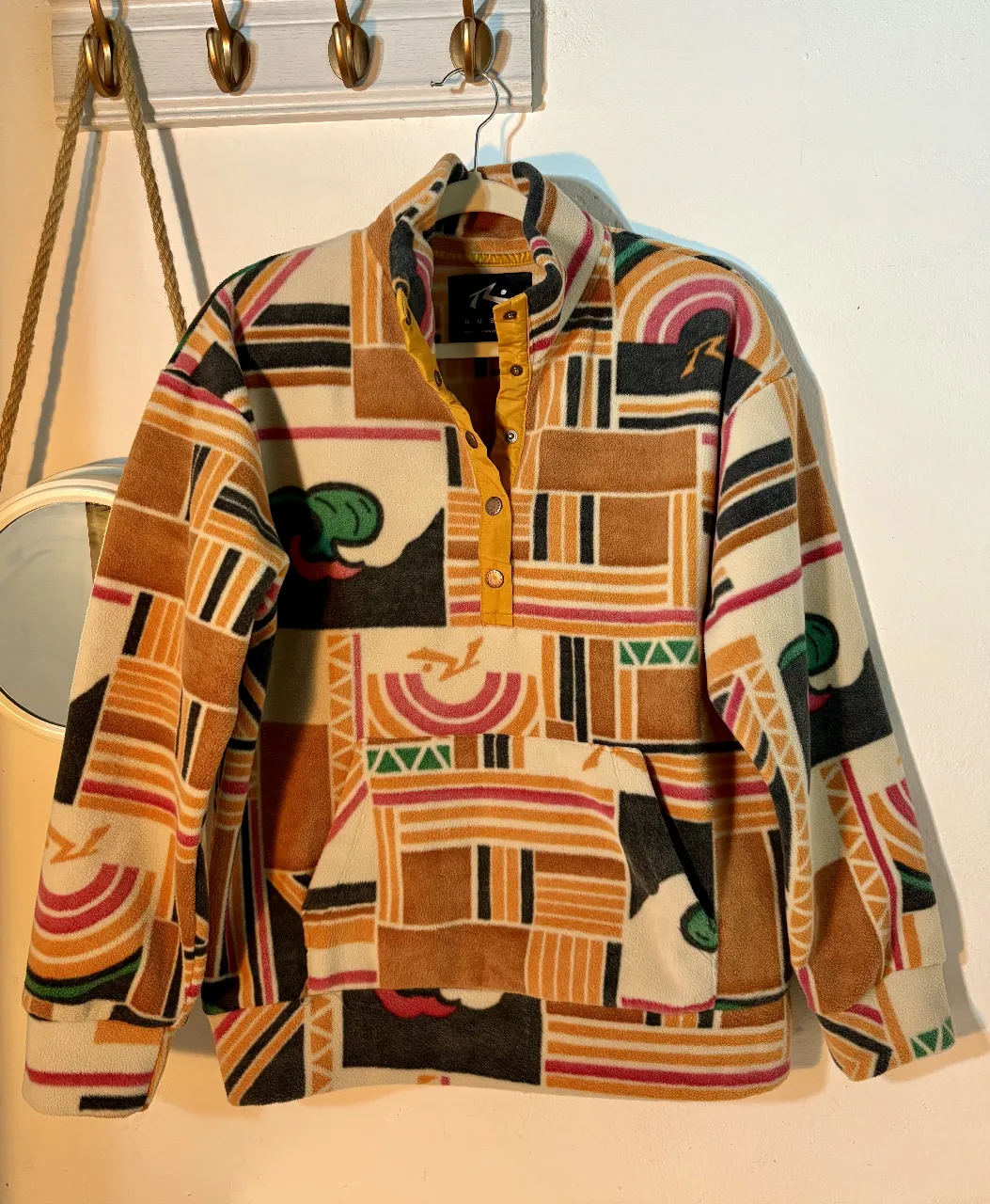 Buzo de polar Rusty con estampado geométrico en tonos marrones, naranjas, verdes y detalles amarillos. Cuello alto con botones y bolsillo frontal. Ideal para un look casual y abrigado.