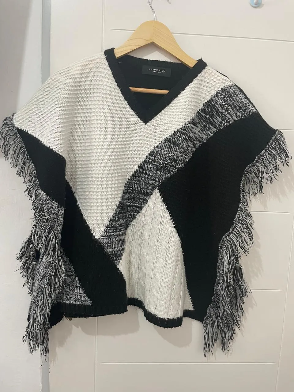 Poncho tejido con escote en V y flecos en los bordes. Diseño geométrico en blanco, negro y gris.