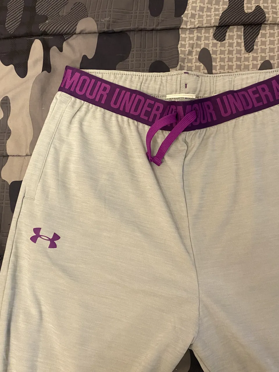 Pantalón deportivo gris claro con cintura elastizada y cordón de ajuste en color violeta. Presenta el logo de la marca en color violeta en la parte superior izquierda.