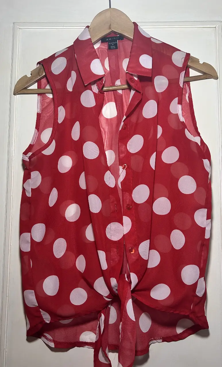 Blusa roja con lunares blancos, sin mangas y con botones. Tiene un lazo en la cintura para ajustar.