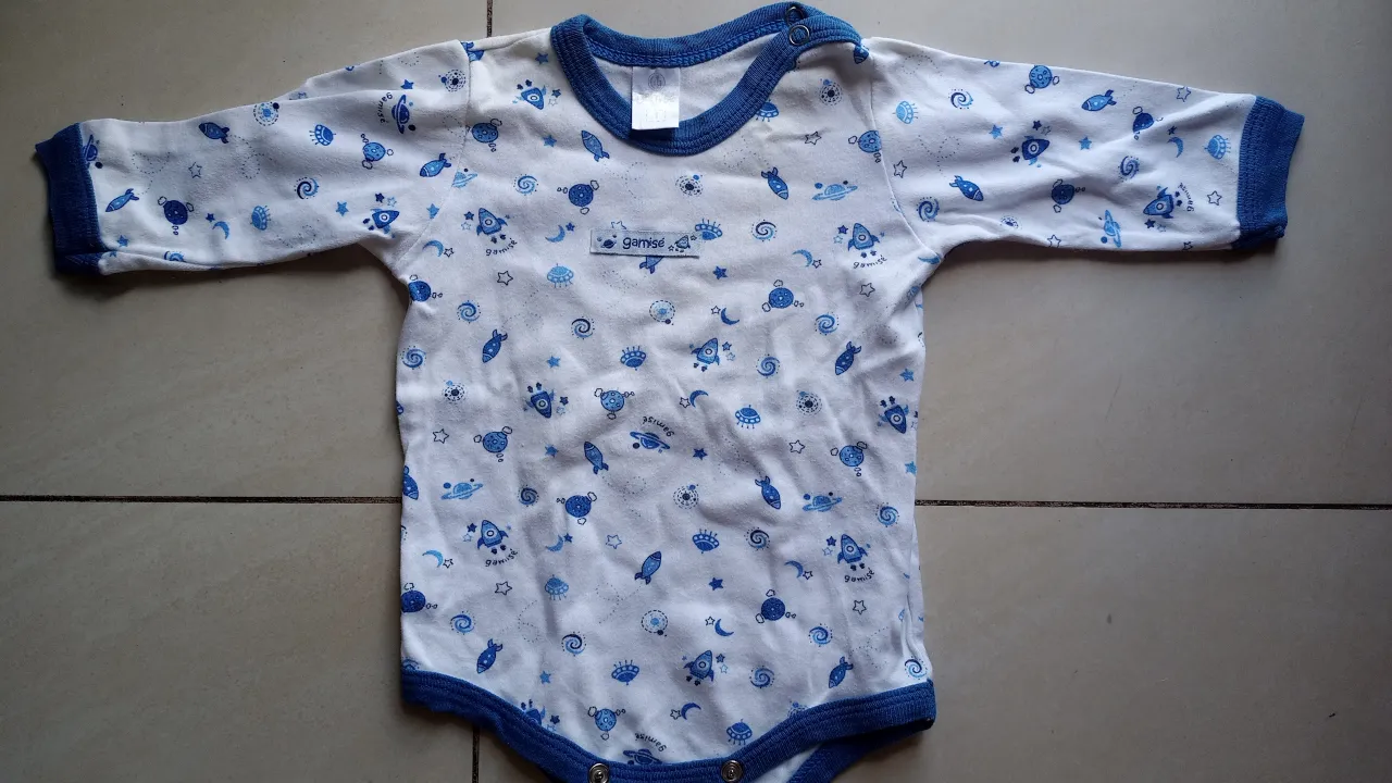 Body de bebé con estampado de cohetes y estrellas en tonos azules sobre fondo blanco. Cuello y puños en azul marino. Ideal para recién nacidos.