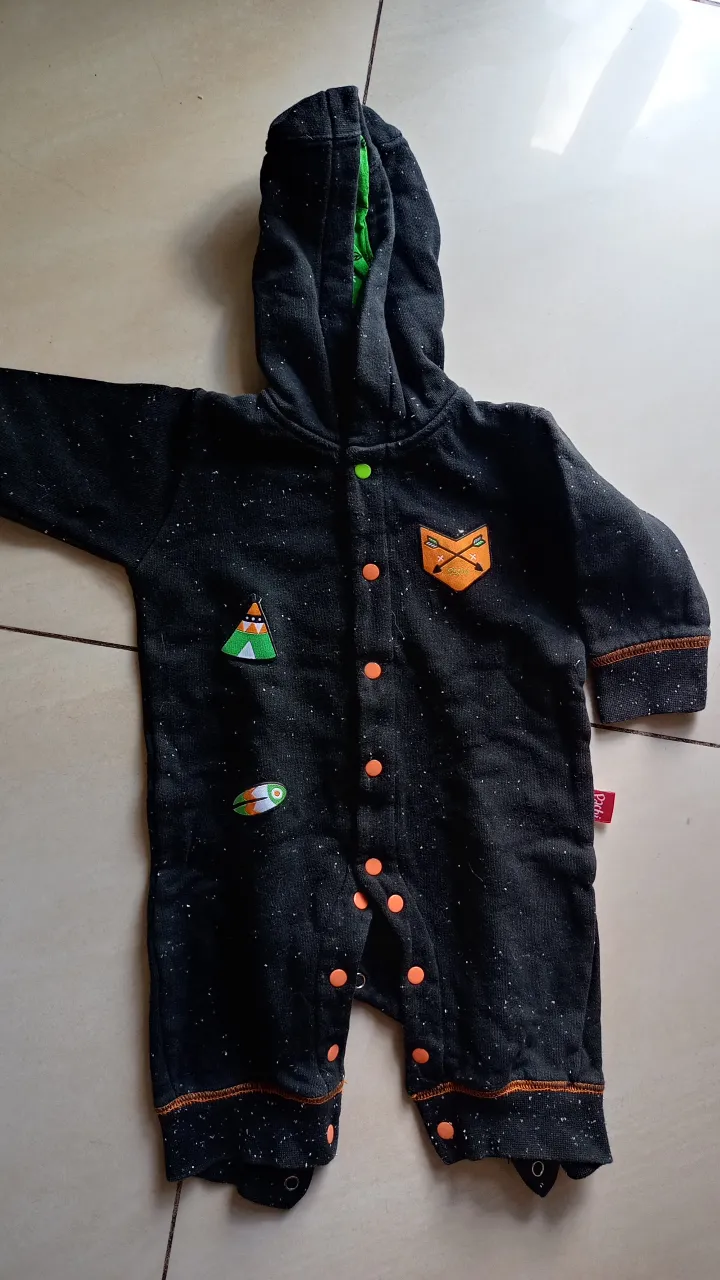 Enterito de bebé con capucha, color negro con detalles en verde y naranja. Diseño con parches decorativos y cierre frontal con botones.