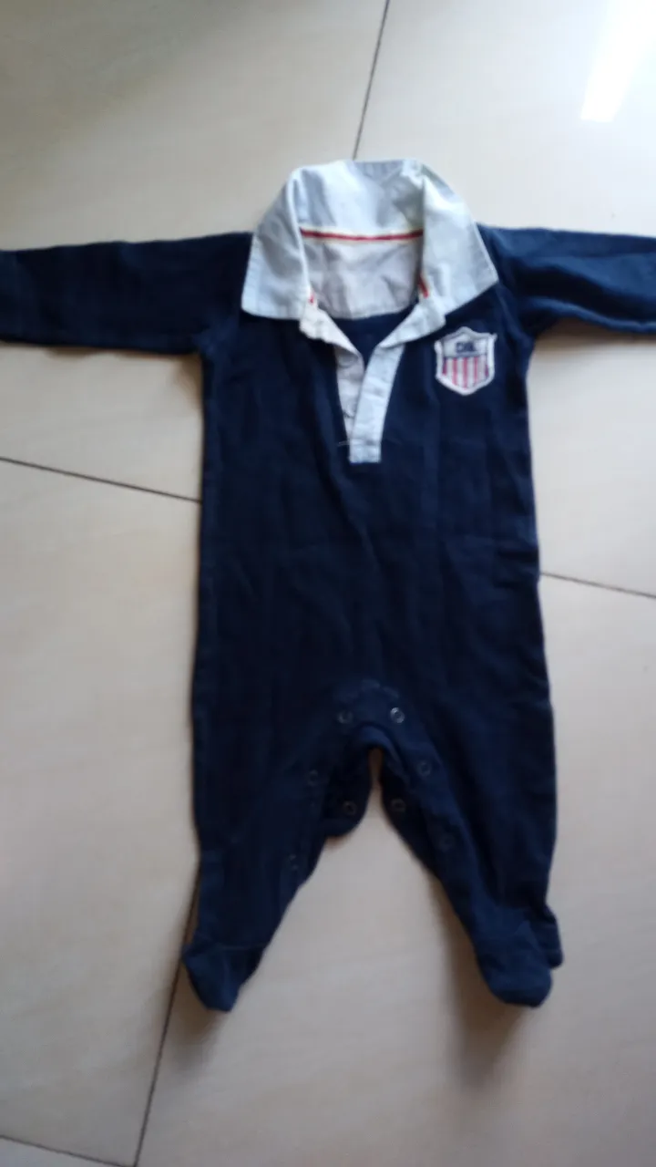 Body de bebé azul marino con cuello blanco y detalles a juego. Diseño con escudo en el pecho y botones en la entrepierna para facilitar el cambio de pañales. Ideal para mantener a tu bebé cómodo y a la moda.