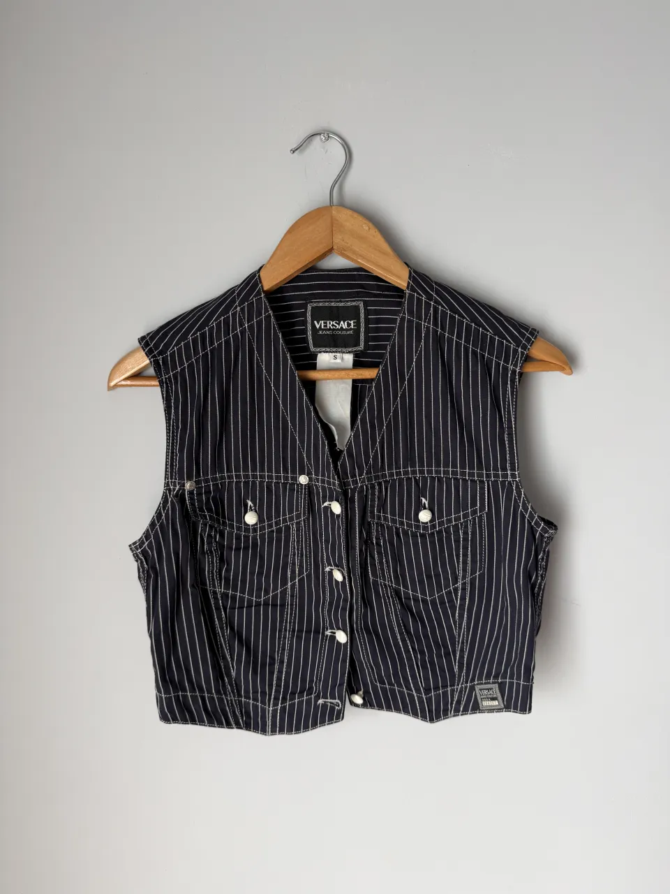 Chaleco  azul oscuro con finas rayas verticales blancas. Posee botones blancos al frente y dos bolsillos con solapa en el pecho. Tiene costuras blancas visibles.