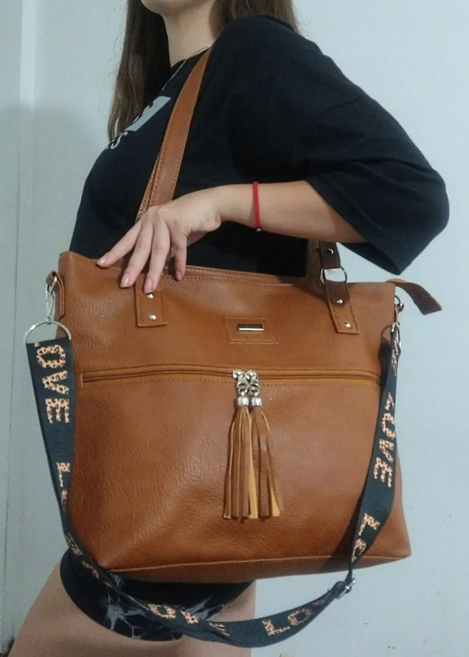 Cartera tipo tote bag de cuero sintético marrón con correa ajustable y desmontable con estampado "LOVE". Posee un bolsillo frontal con cierre y detalles de borlas.