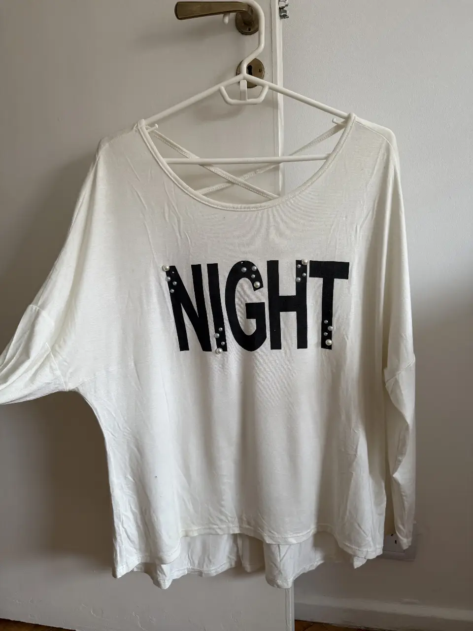 Remera manga larga blanca con estampado "NIGHT" en negro y detalles de tachas. Diseño con espalda cruzada y cuello redondo. Ideal para un look casual y moderno. Tiene una pequeña mancha en la parte inferior delantera