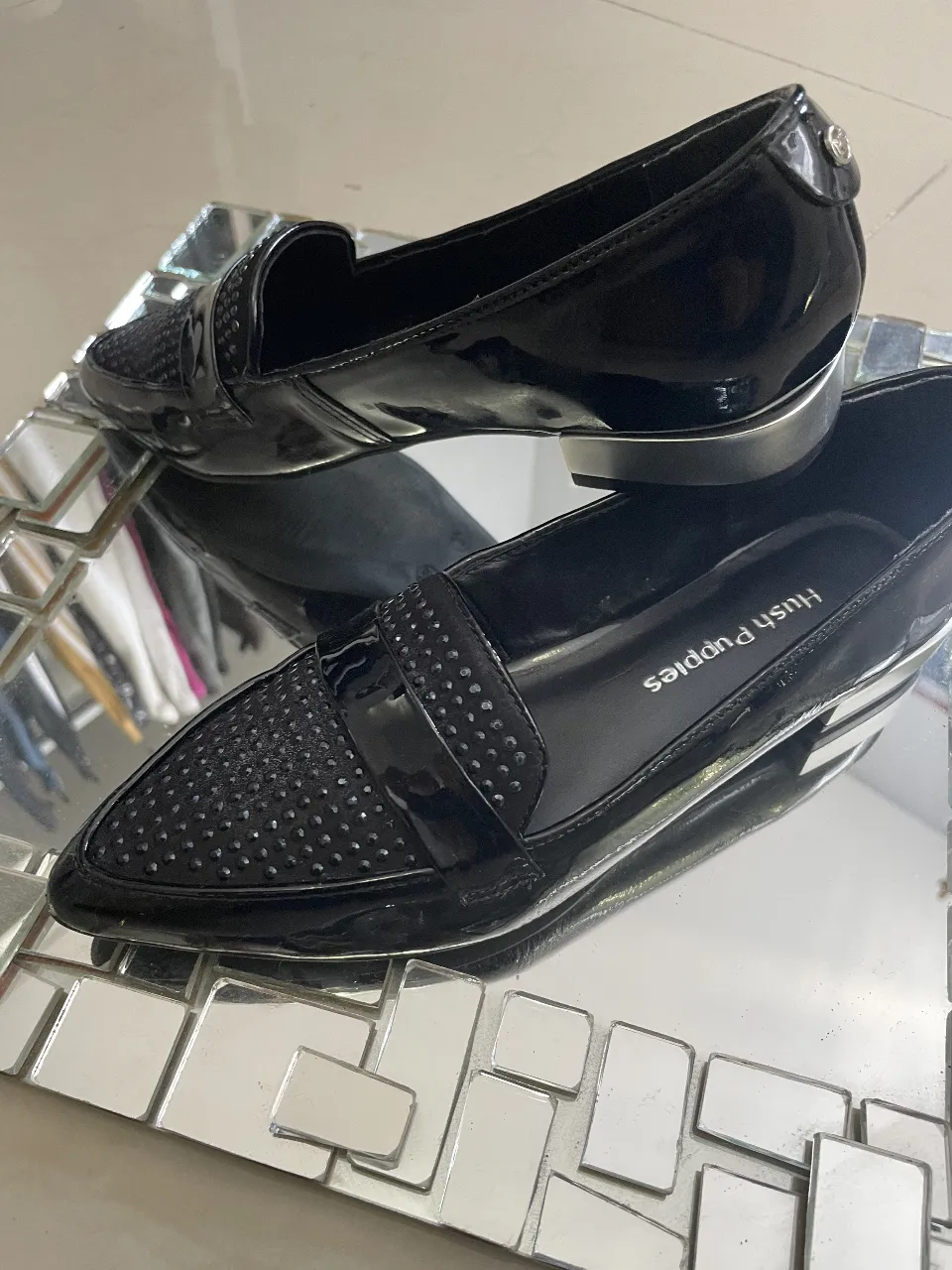 Elegantes mocasines Hush Puppies para mujer, en charol negro con detalles brillantes en la puntera. Diseño sofisticado con suela plateada, ideales para un look de fiesta o salida.