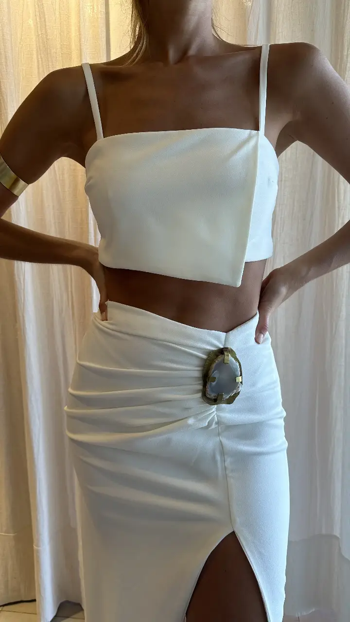Conjunto de novia de dos piezas blanco. Tela de crep. El top, con diseño asimétrico y tirantes finos, realza la figura. La falda, de corte midi con abertura lateral, presenta un detalle drapeado y un aplique de piedra en una base de bronce. marca Inejo