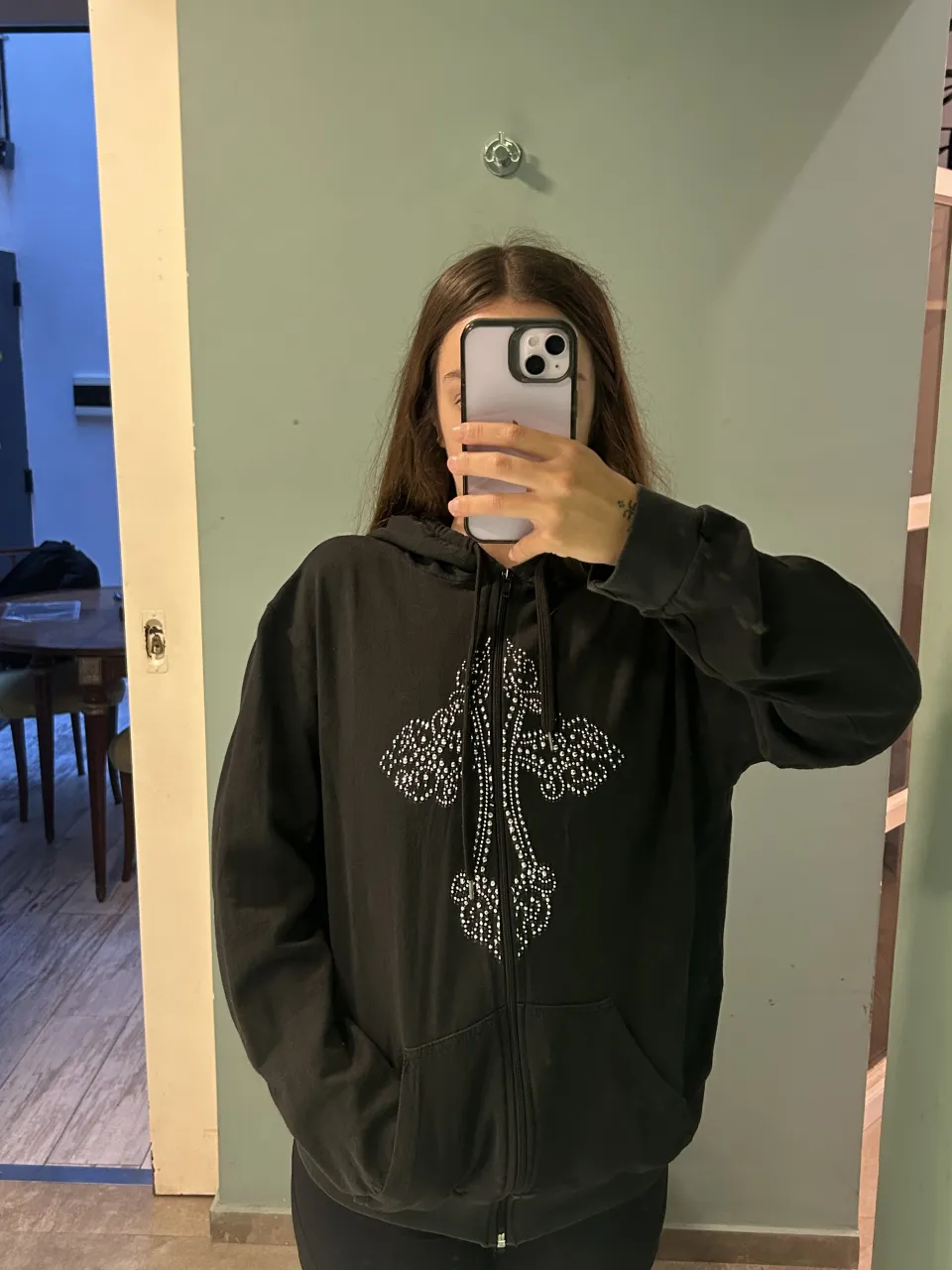 Buzo negro con cierre y capucha, tiene un diseño de cruz con pedrería blanca en el frente. Posee bolsillos tipo canguro.