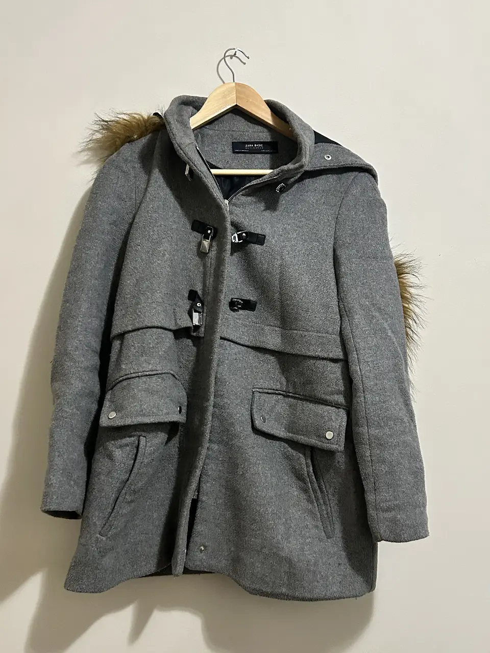 Tapado gris de paño con capucha y detalles de piel sintética en la capucha. Posee bolsillos delanteros y cierre con broches. Ideal para el invierno.