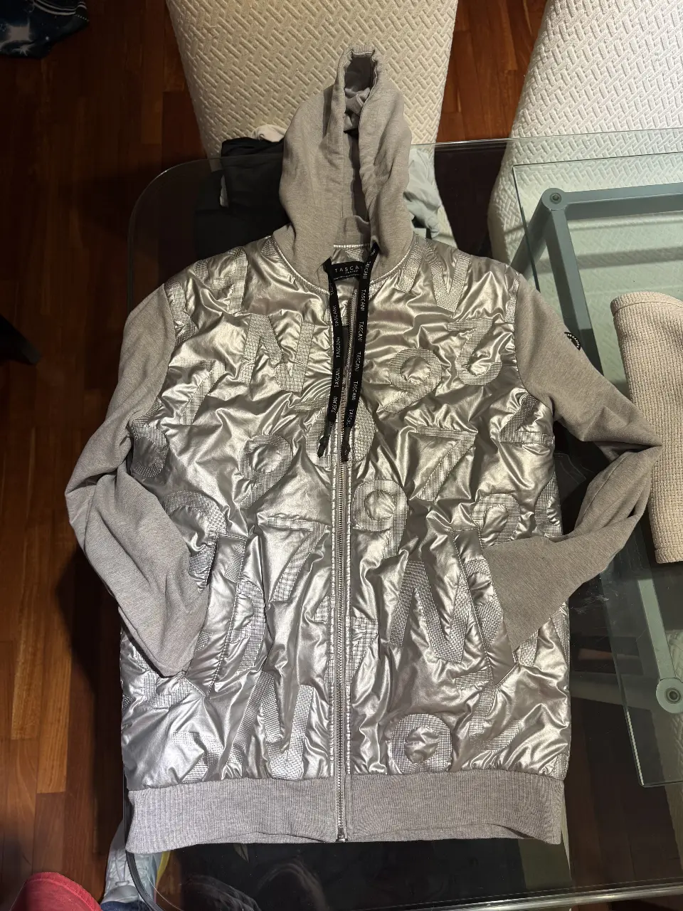 Buzo con cierre para hombre, de la marca Tascani. Confeccionado con un diseño acolchado plateado en la parte delantera y mangas grises. Posee capucha y cierre frontal. Ideal para un look casual y moderno. excelente estado 
