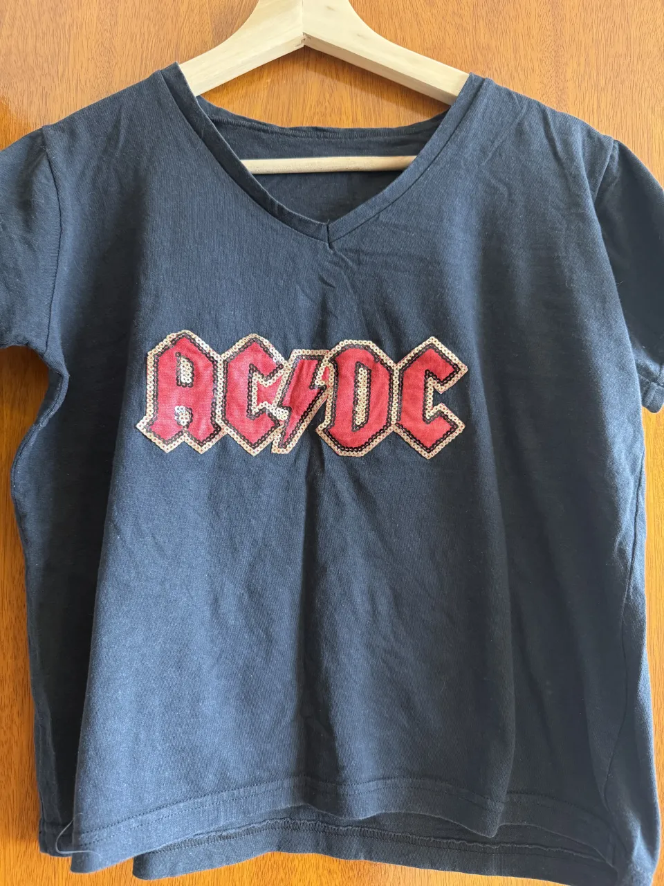 Remera negra de manga corta con cuello en V. Presenta un estampado de lentejuelas rojas y doradas con el logo de AC/DC.