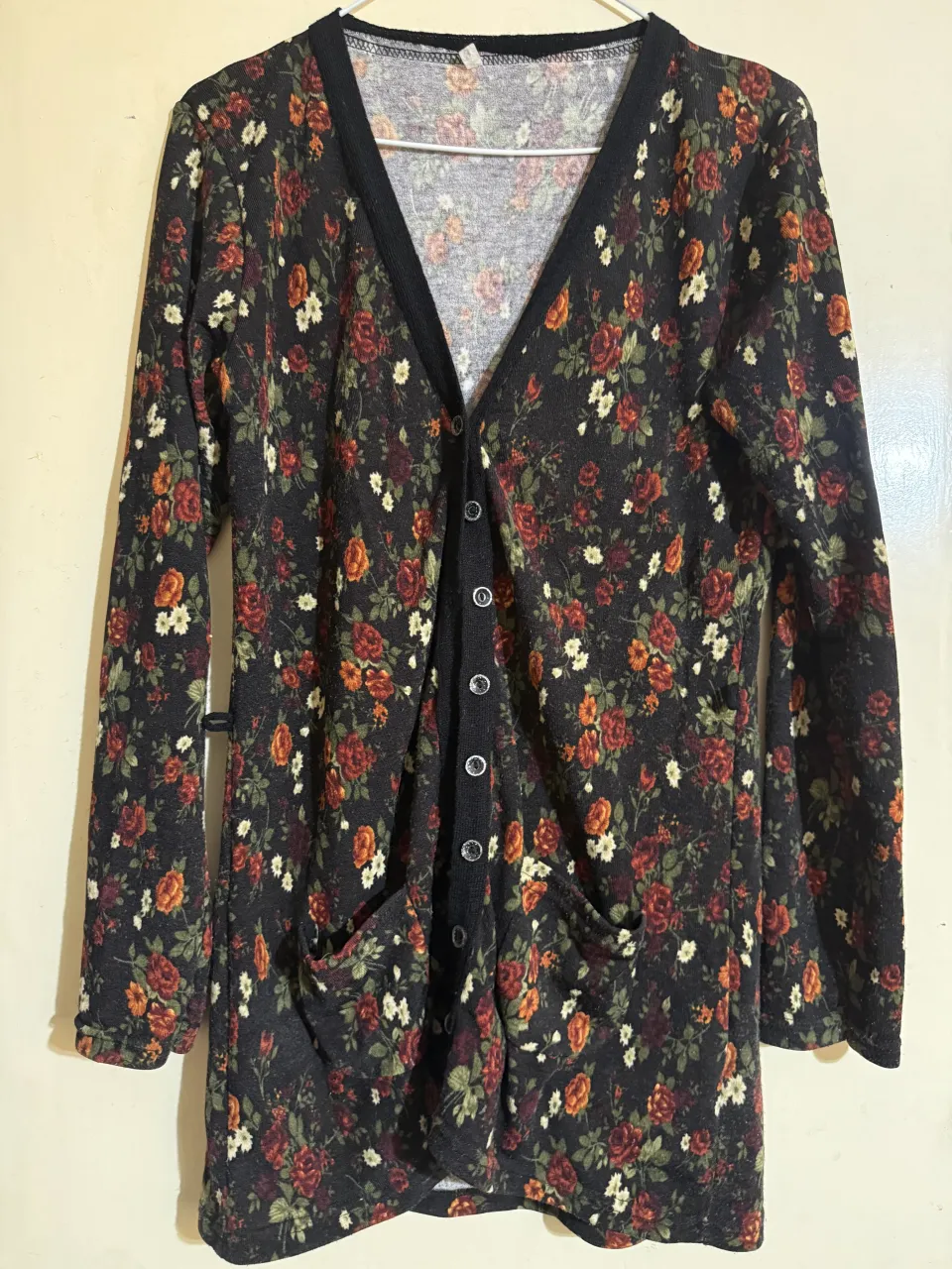 Cardigan negro con estampa floral de rosas en tonos rojos, naranjas y blancos. Posee cuello en V, botones al frente y dos bolsillos aplicados.
