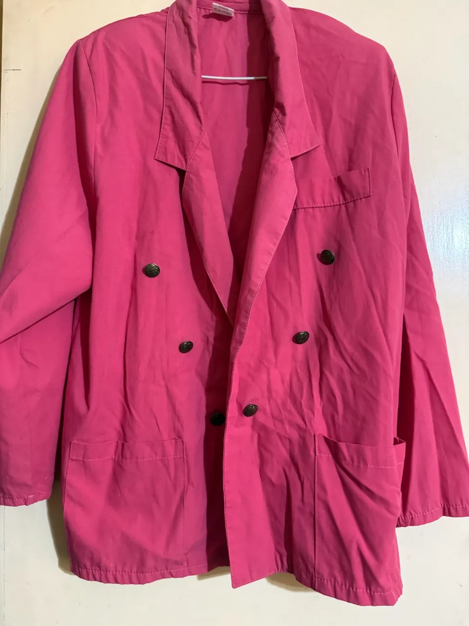 Blazer rosa de botones dorados. Tiene dos bolsillos delanteros y un bolsillo superior.