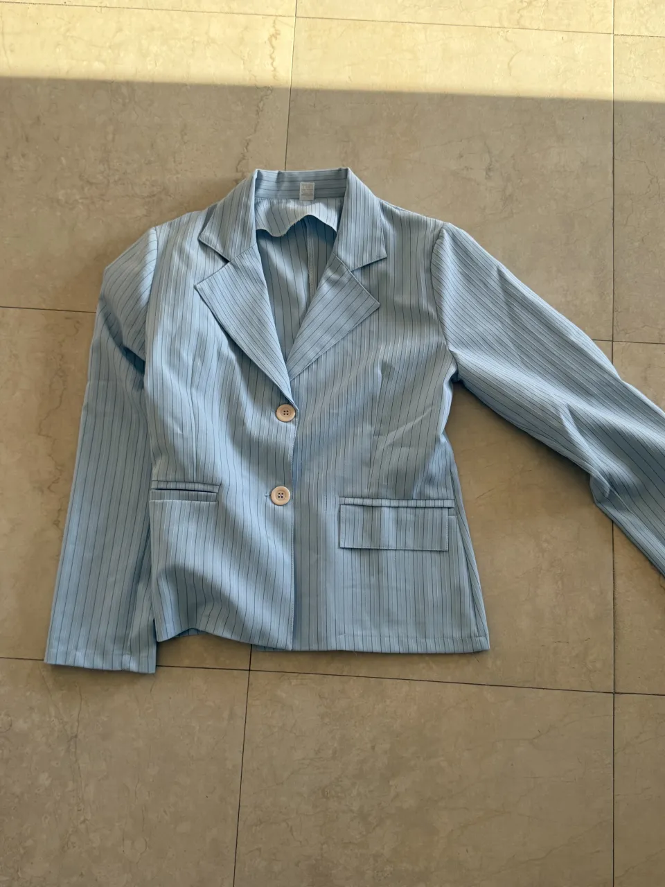 Blazer celeste con rayas verticales finas y dos botones al frente. Posee bolsillos con solapa.