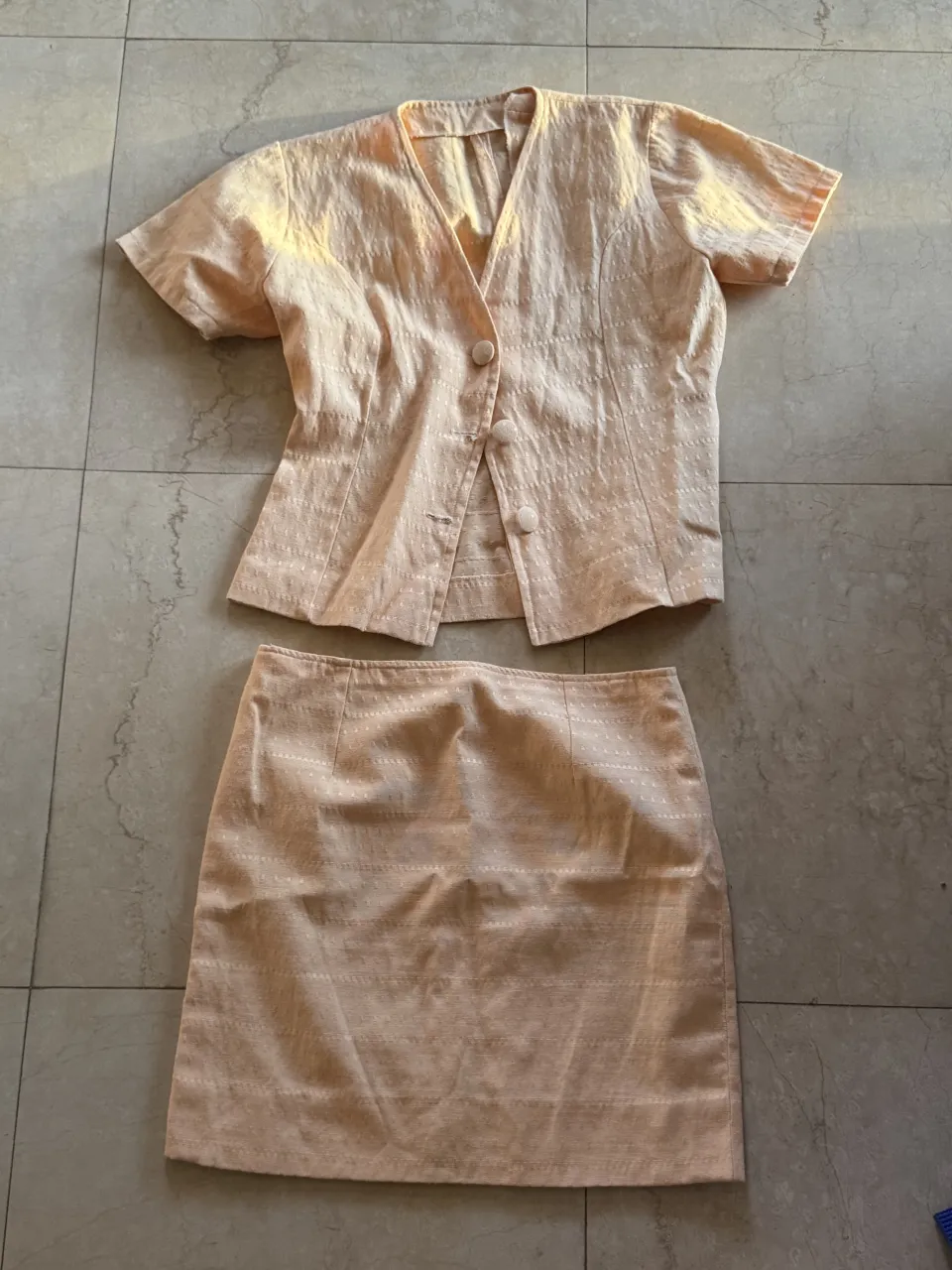 Conjunto de blusa y pollera color salmón. La blusa es de manga corta, con cuello en V y tres botones al frente. La pollera es recta y de largo hasta la rodilla.