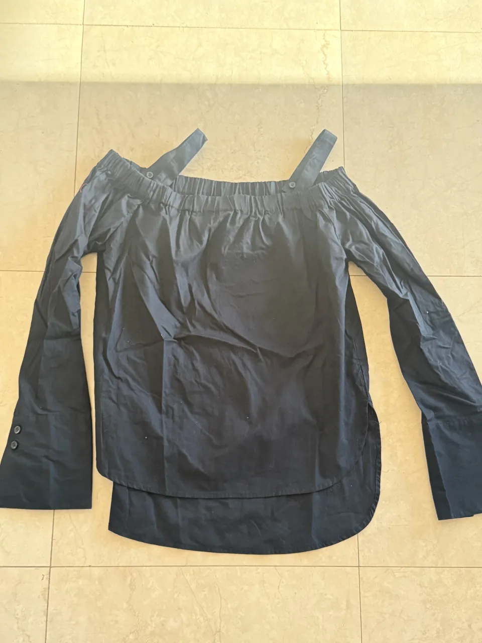Blusa negra de mangas largas con hombros descubiertos y tiras. Posee botones en los puños y un ruedo curvo.