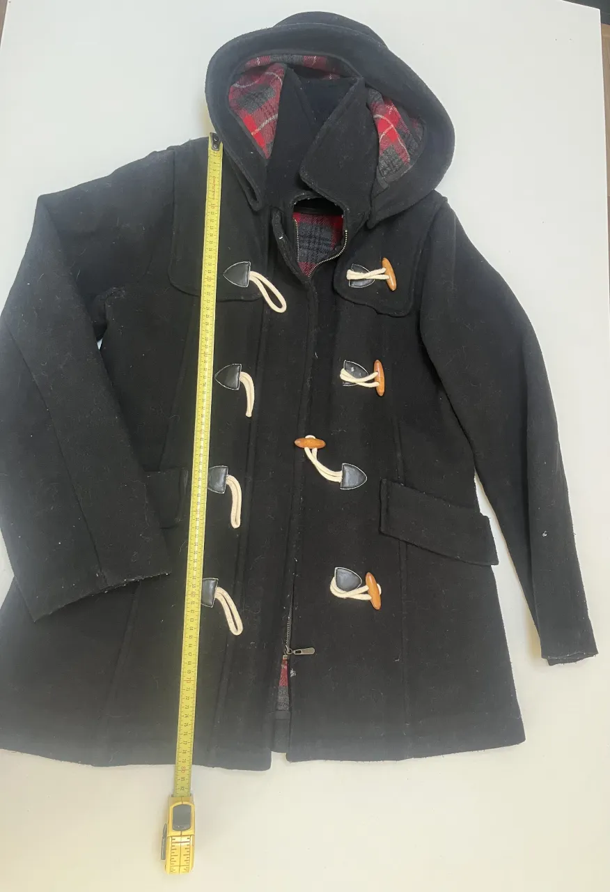 Abrigo negro estilo Duffle Coat con detalles de cuero y forro a cuadros escoceses. Posee cierre y botones de madera y soga. Ideal para un look casual y abrigado.