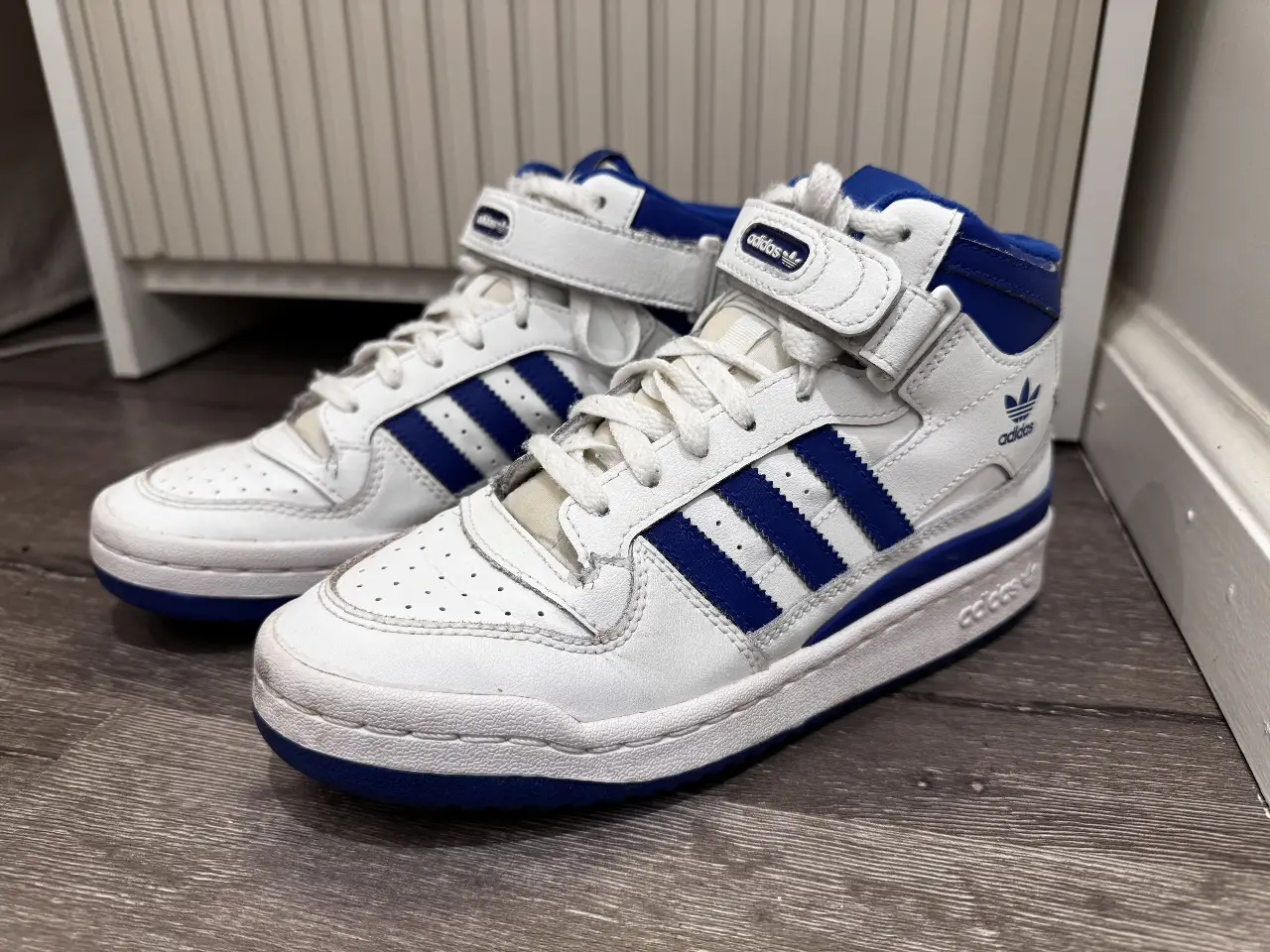 Zapatillas Adidas Forum Mid blancas con detalles en azul. Diseño icónico de caña alta con cierre de velcro y las clásicas tres tiras. Ideales para un look urbano y deportivo.