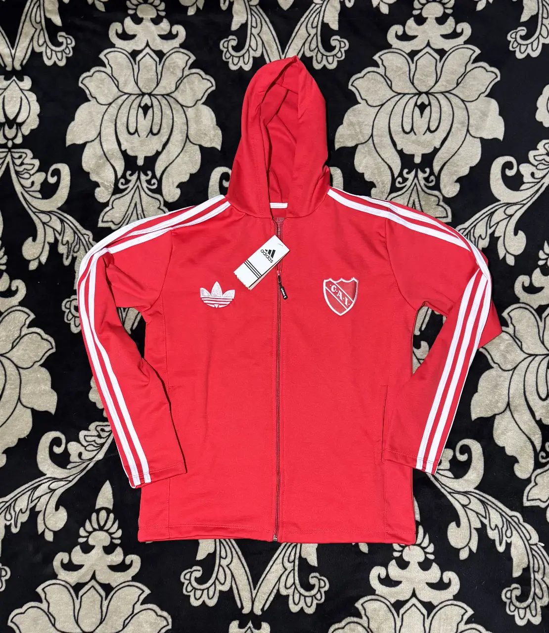 Buzo deportivo con cierre y capucha, de color rojo con detalles blancos. Presenta el logo de Adidas y el escudo del club. Ideal para los fanáticos del fútbol y para un look casual y deportivo. Talles del M al XXL