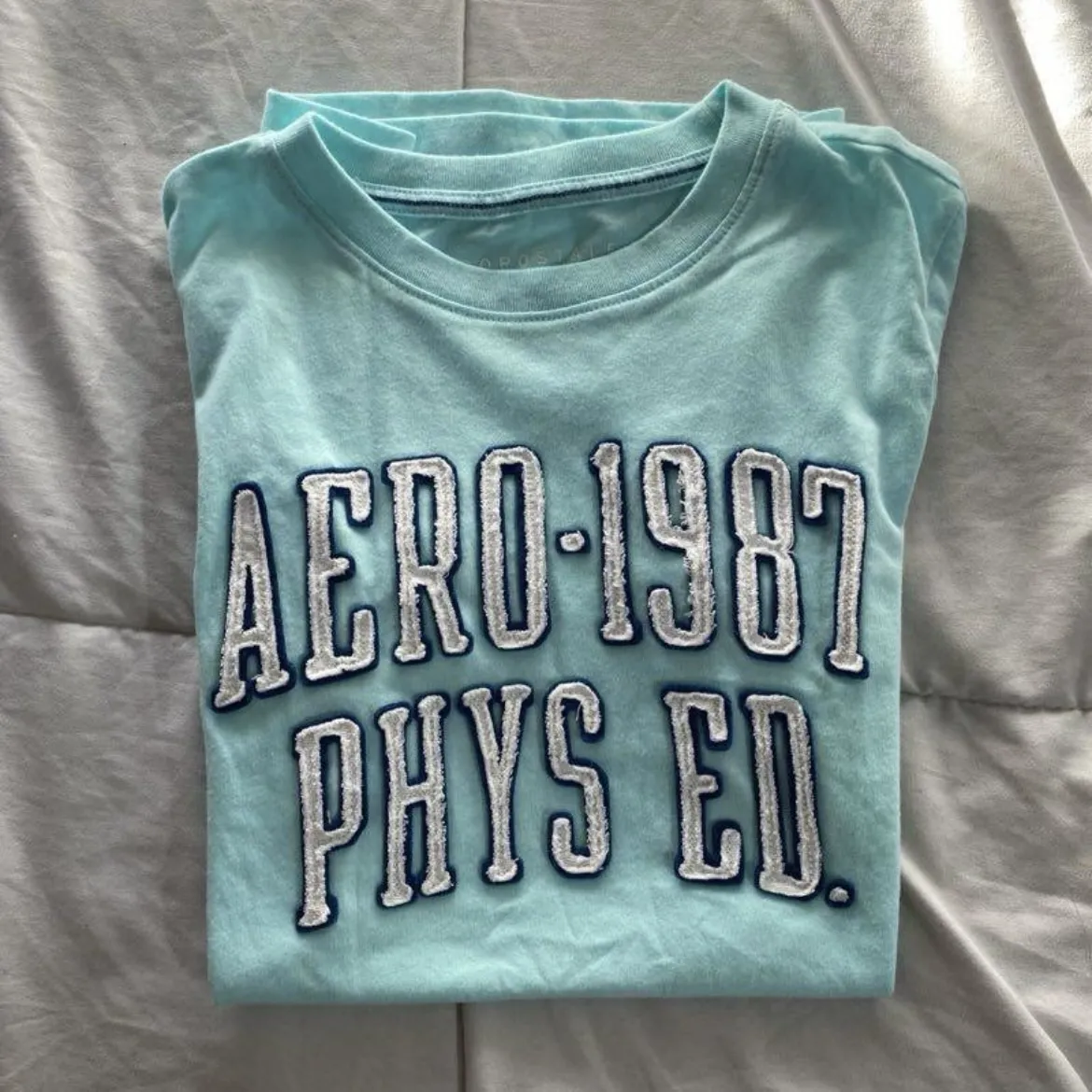 Remera deportiva celeste de manga corta con estampado "AERO-1987 PHYS ED". Ideal para actividades físicas o uso casual.