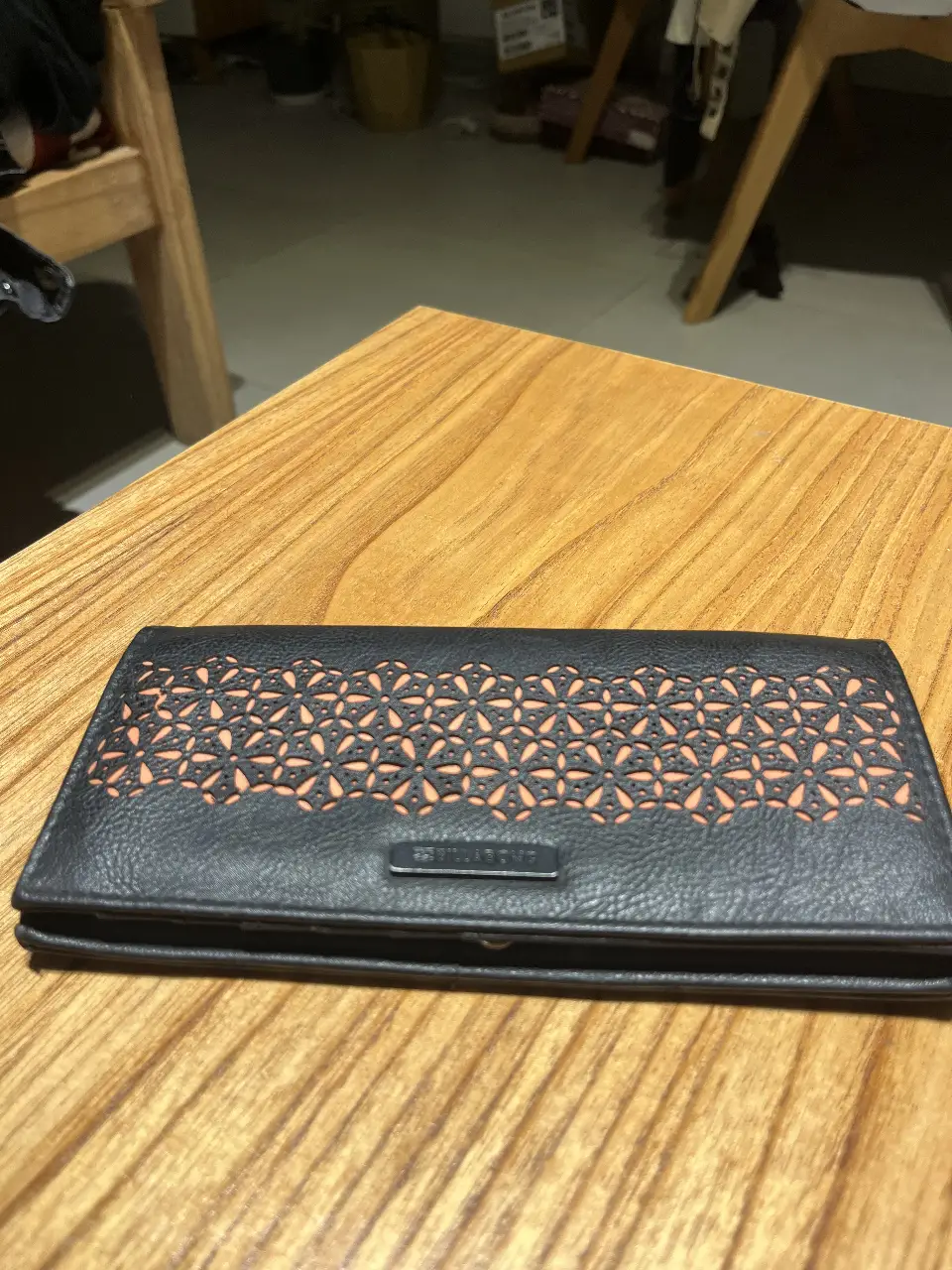 Billetera de cuero negro con diseño calado en la parte superior y detalles en naranja. Cuenta con múltiples compartimentos para tarjetas y billetes, y un cierre de botón.