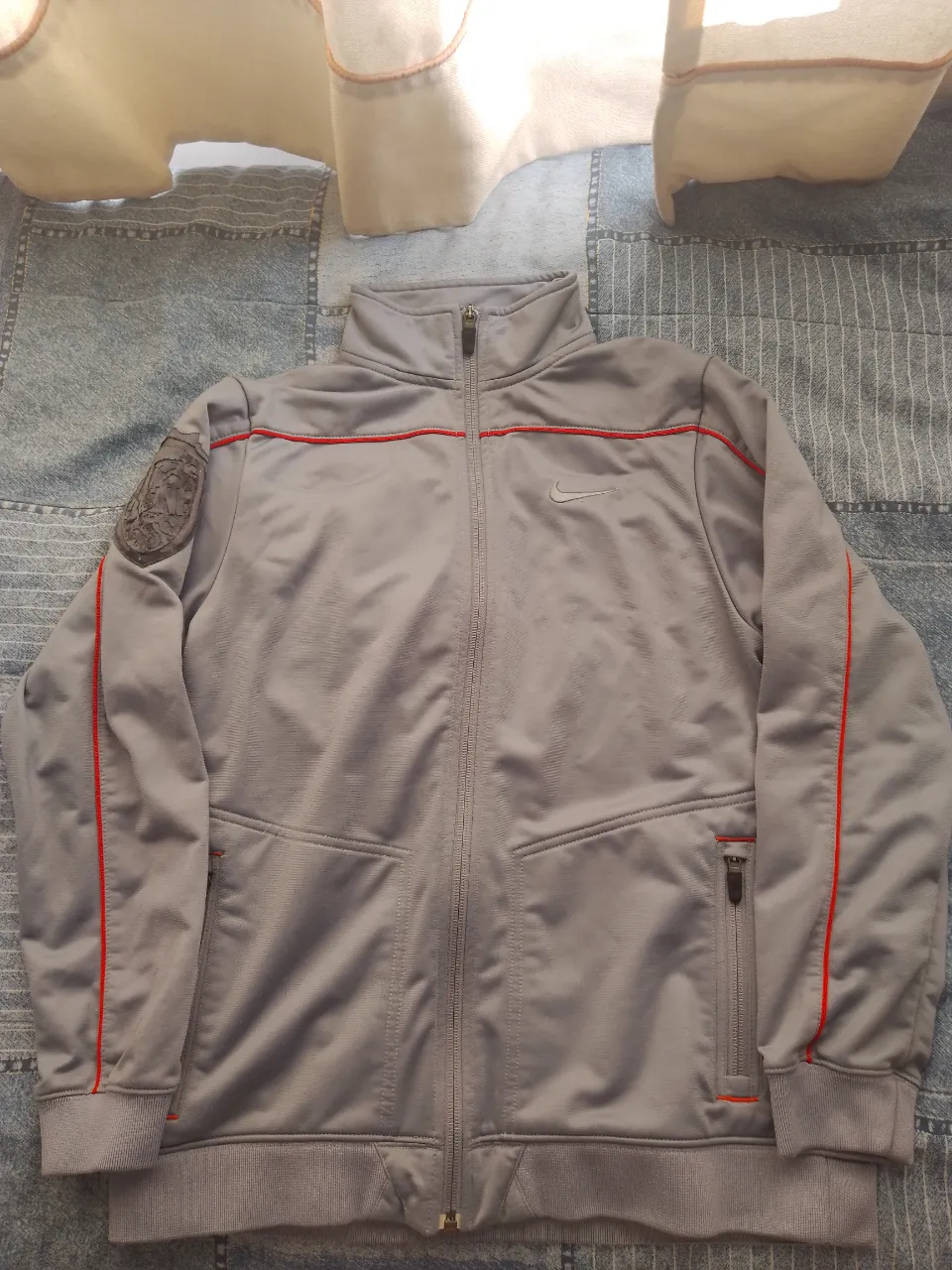 Buzo deportivo gris con detalles en rojo, cierre completo y bolsillos con cierre. Ideal para entrenamientos o uso casual.