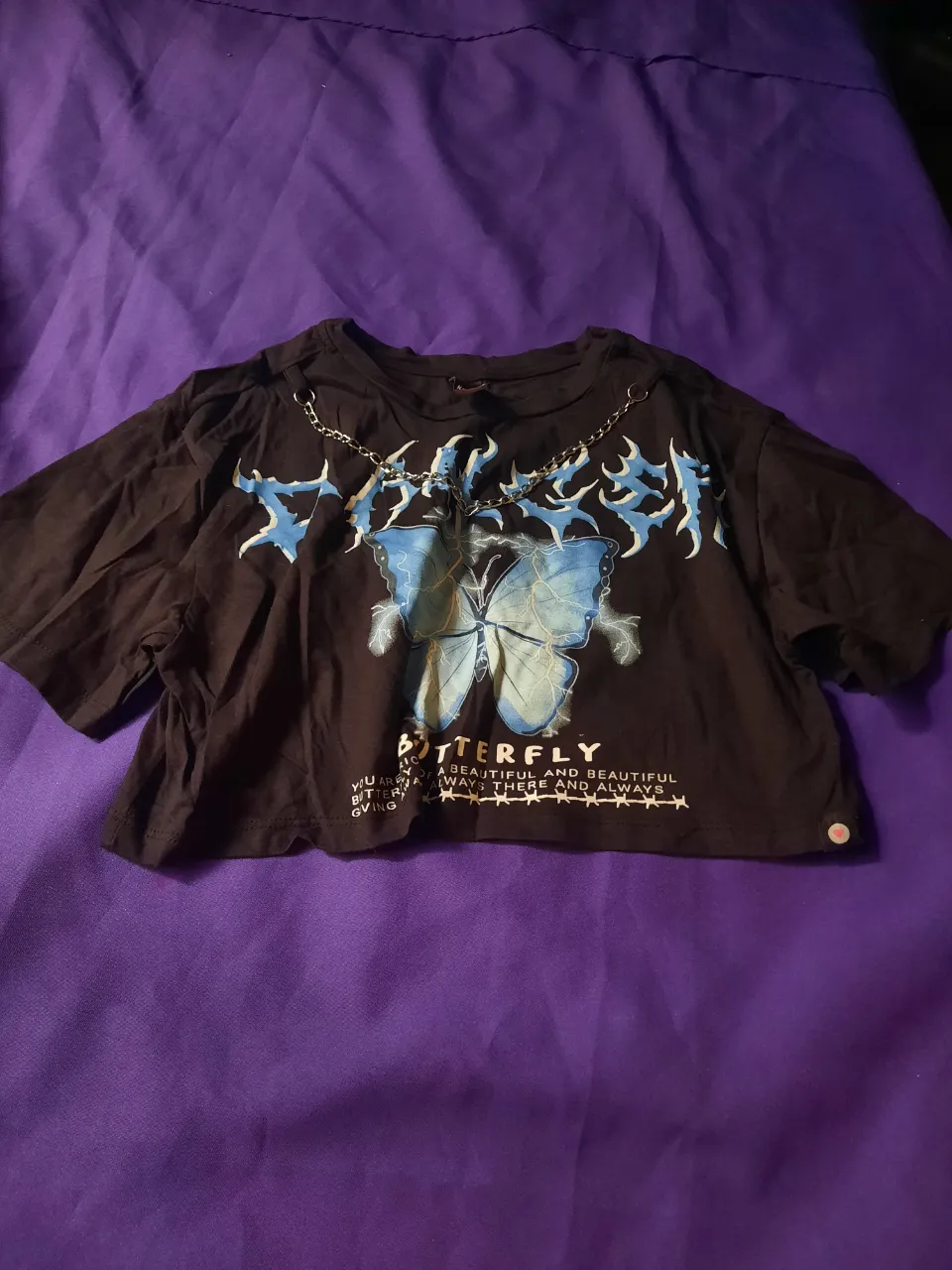 Remera negra crop con estampa de mariposa y cadenas. Tiene inscripciones en la parte inferior. Talle S/M.