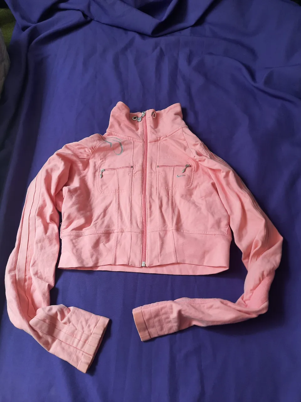 Campera deportiva rosa, corte crop, con cuello alto y cierre al frente. Posee bolsillos con cierre y un diseño estampado en la espalda. Ideal para un look casual y deportivo.