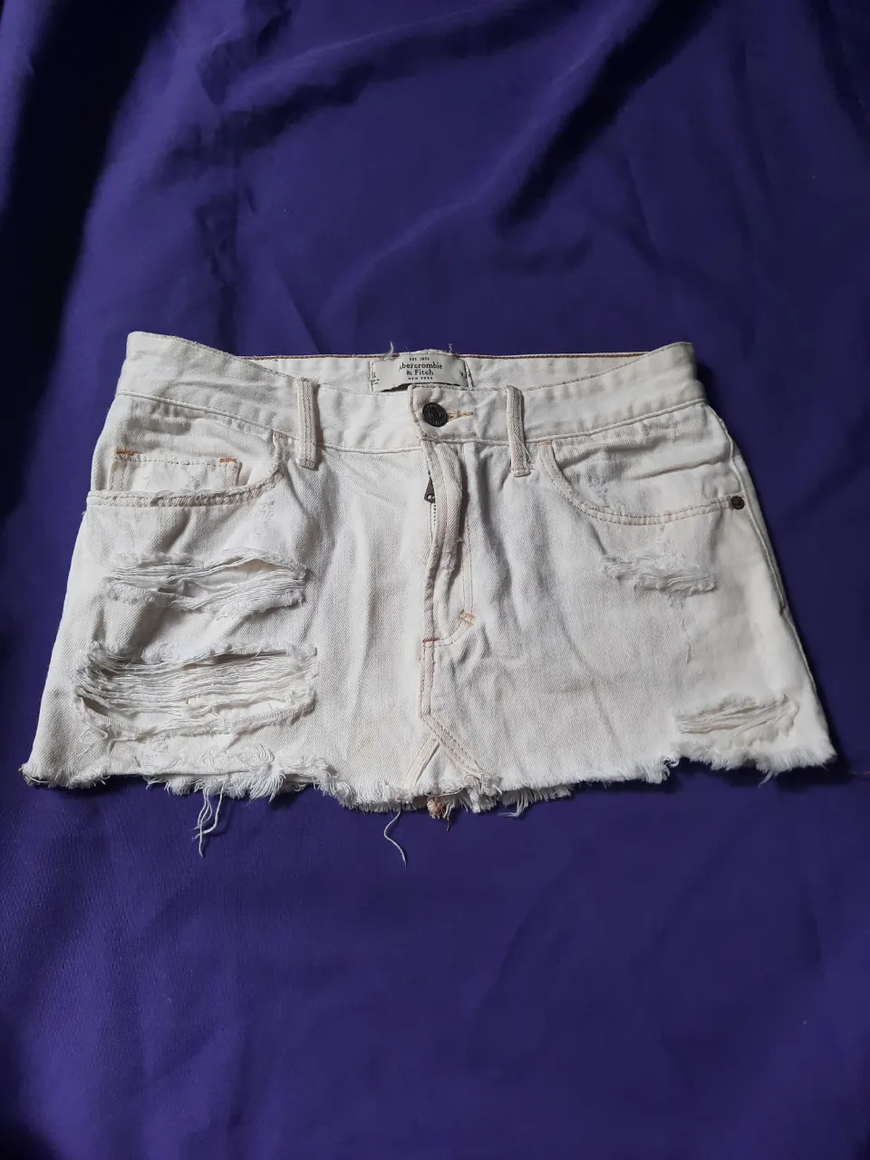 Pollera de jean tiro bajo blanca con roturas y detalles deshilachados en el borde inferior. Tiene bolsillos delanteros y traseros.