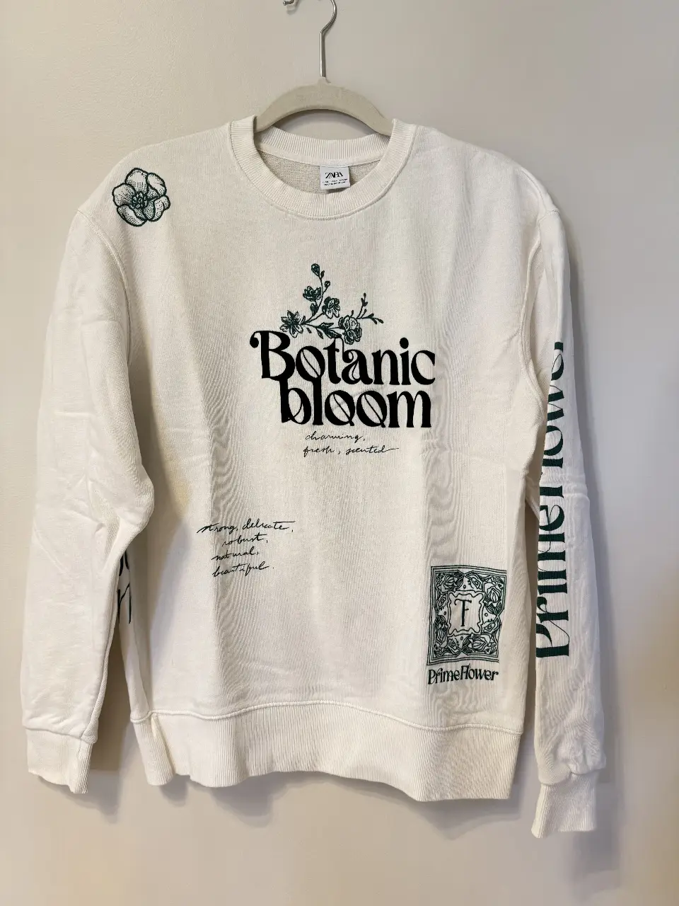 Buzo de cuello redondo de algodón color blanco con estampado "Botanic Bloom" en verde. Diseño con detalles florales y frases en la manga y en la parte delantera y trasera. Ideal para un look casual y cómodo.