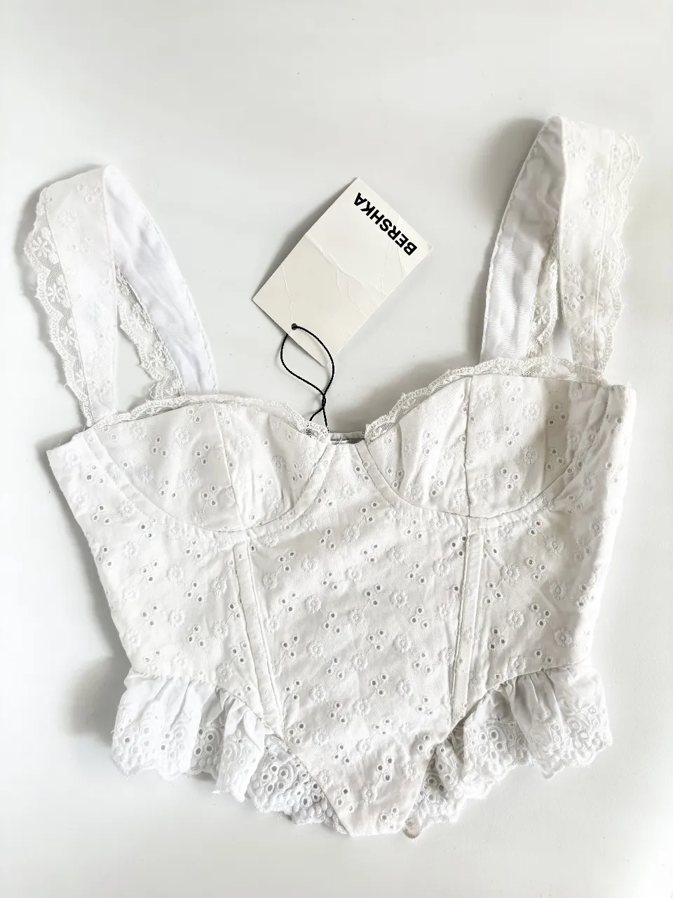 Top blanco de Bershka con bordado floral y detalles de encaje. Tiene tirantes anchos y un diseño tipo corset con volados en la parte inferior. Cuenta con un cierre escondido al costado