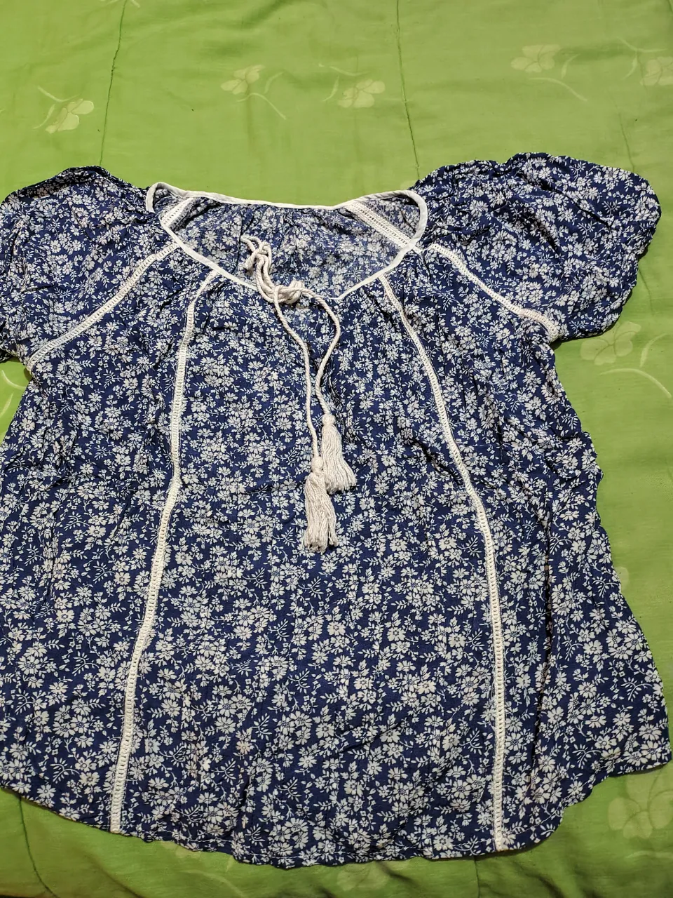 Blusa estampada con flores azules y blancas, escote en V con cordones y pompones, mangas cortas y detalles de puntilla blanca. Ideal para un look fresco y bohemio.