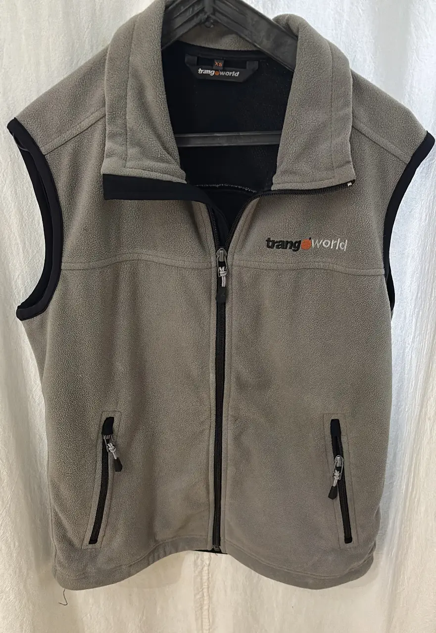 Chaleco gris de polar con cierre frontal y dos bolsillos con cierre. Tiene el logo de la marca "trangoworld" bordado en naranja. Ideal para usar en actividades al aire libre o como una capa adicional de abrigo. Marca de ropa técnica para deportes extremos europea