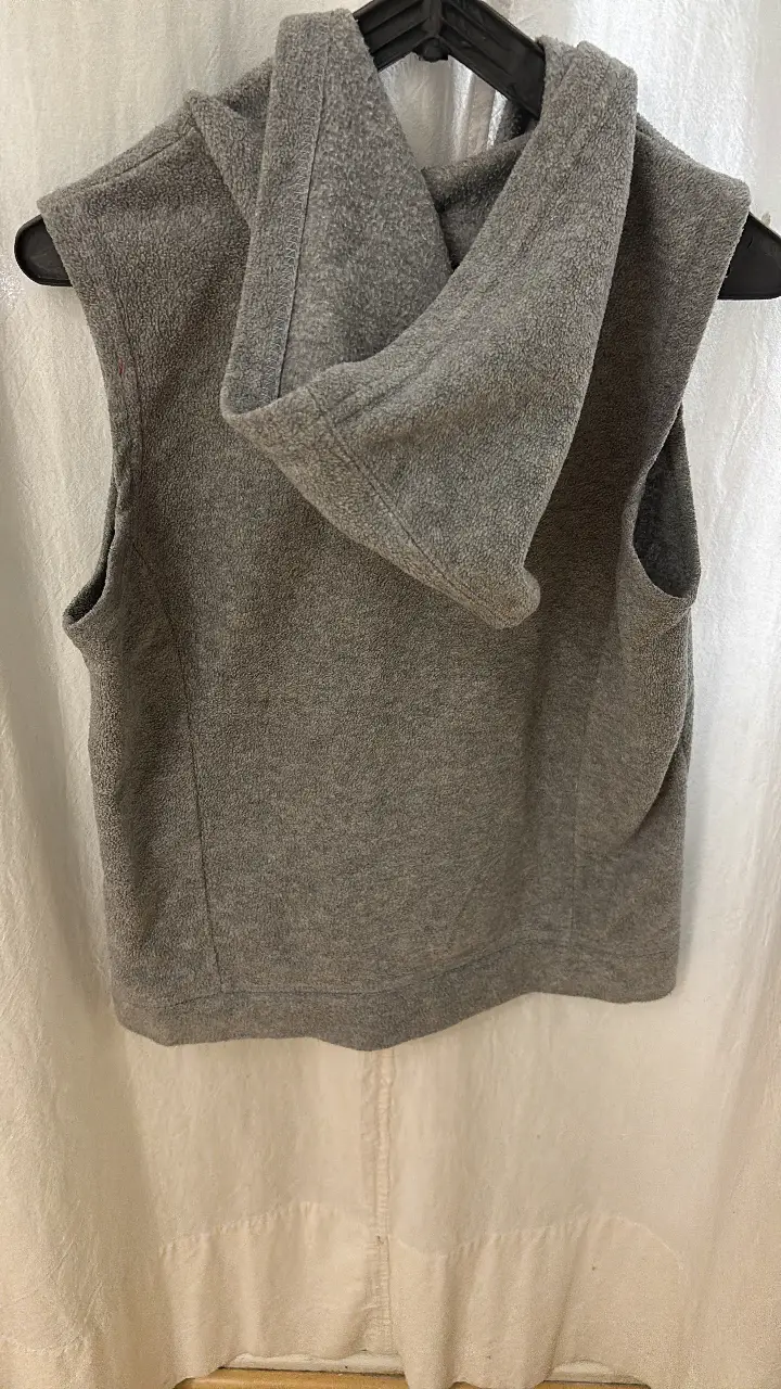 Chaleco con capucha de polar gris, sin mangas, con cierre frontal y bolsillos. Ideal para usar en días frescos.