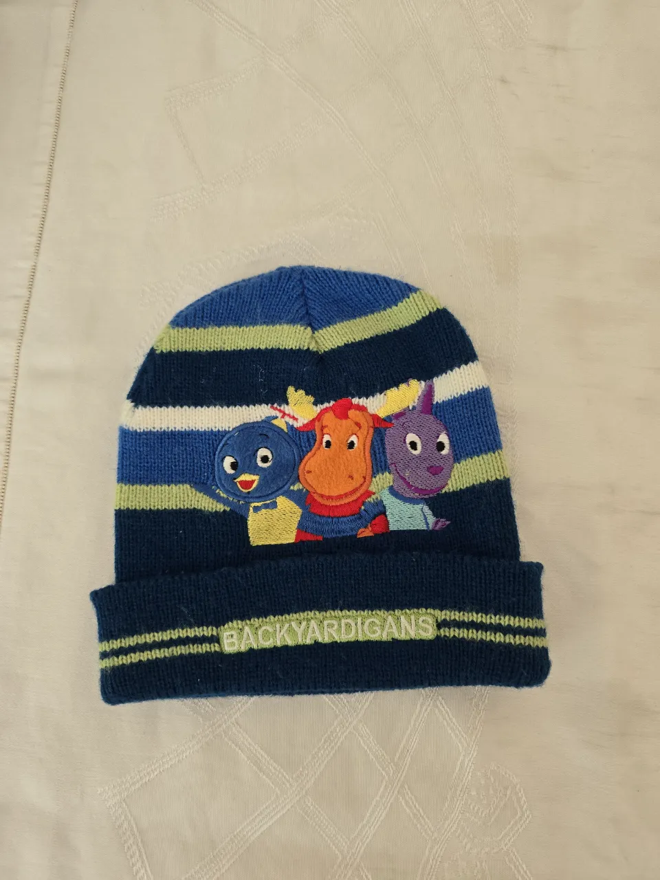 Gorro de lana azul con rayas horizontales de colores verde, blanco y azul oscuro. En el frente, tiene un bordado de tres personajes animados y la inscripción "BACKYARDIGANS".