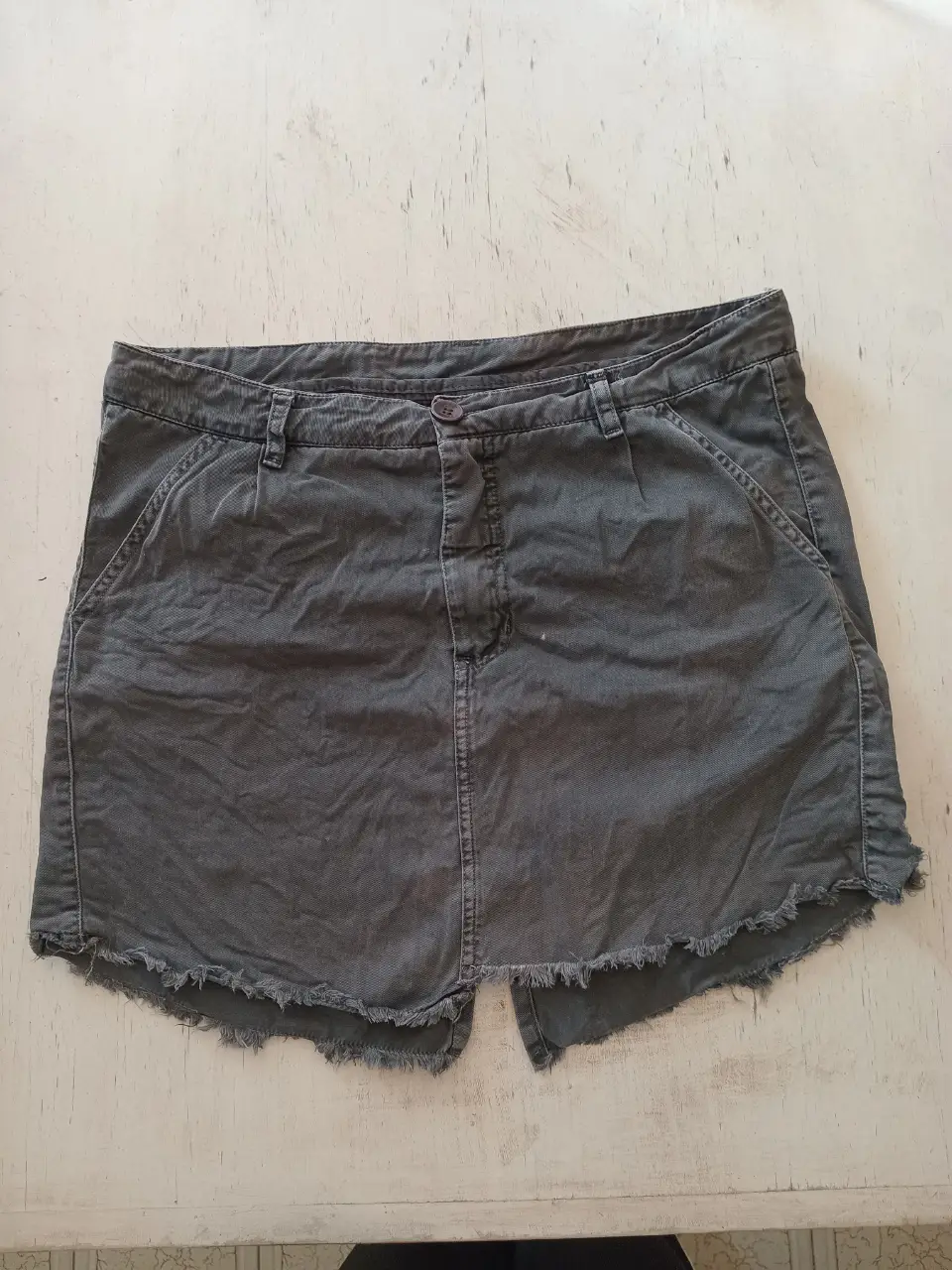 falda de gabardina gris con detalles desflecados en el borde inferior. Posee bolsillos delanteros y traseros, y cierre con botón y cremallera. Ideal para un look casual y cómodo. Detalle costura arreglada lado izquierdo trasero