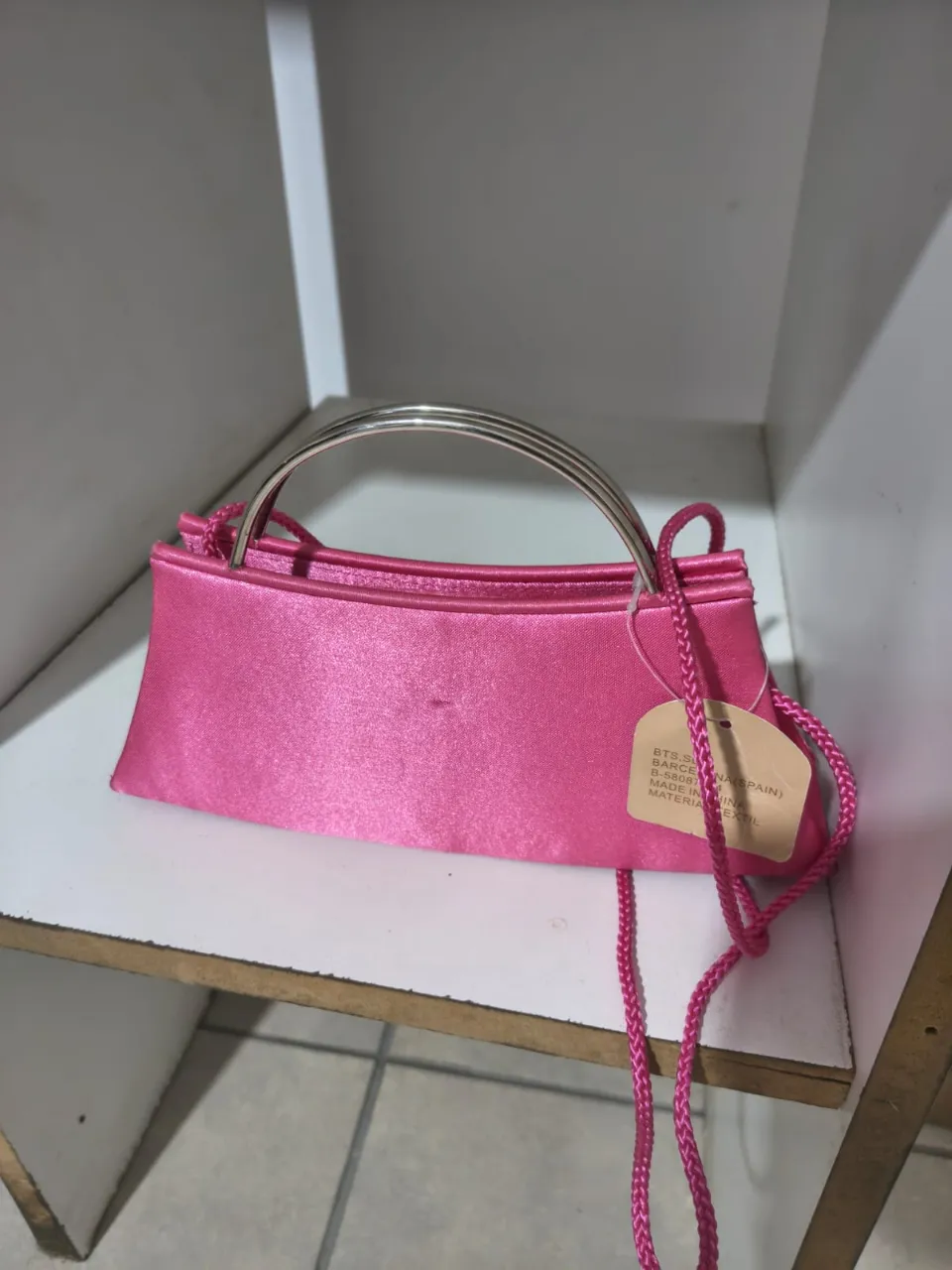 Cartera de mano color rosa fuerte con manija metálica y correa larga de tela. Cierre superior.