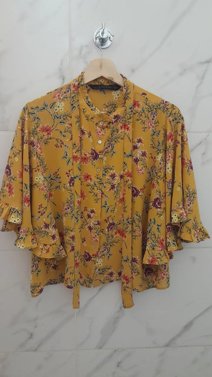 Camisa amarilla con estampa floral vibrante y detalles de volados en mangas y cuello. Posee lazo para atar en el cuello. Ideal para un look fresco y femenino.