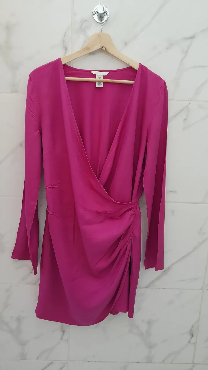 Vestido rosa fucsia con mangas largas y escote cruzado. Tiene un detalle de fruncido en la cintura y falda asimétrica. Ideal para una salida o fiesta.
