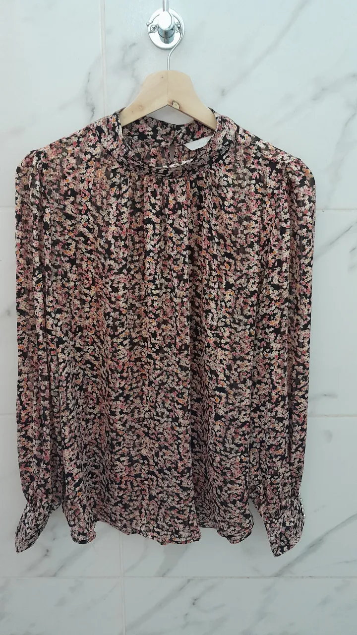 Blusa de gasa estampada con flores pequeñas en tonos rosas, naranjas y negros. Posee cuello alto con fruncido y puños con botones. Ideal para un look elegante y femenino.